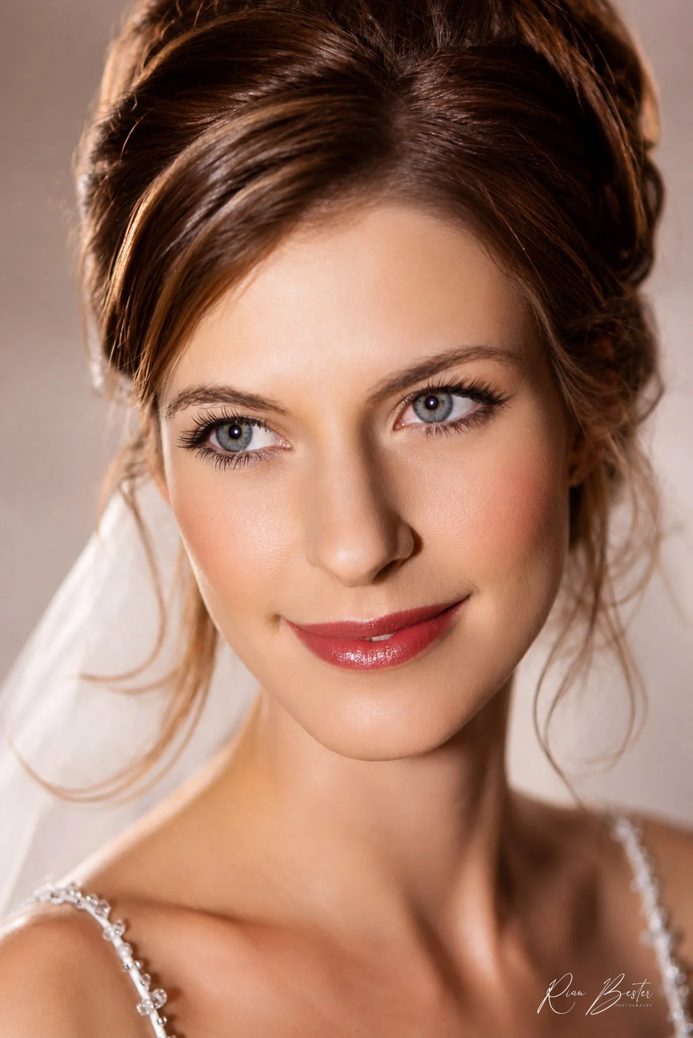 Close-up of a woman with blue eyes, brown hair styled back, wearing a wedding dress with beaded straps, and smiling.