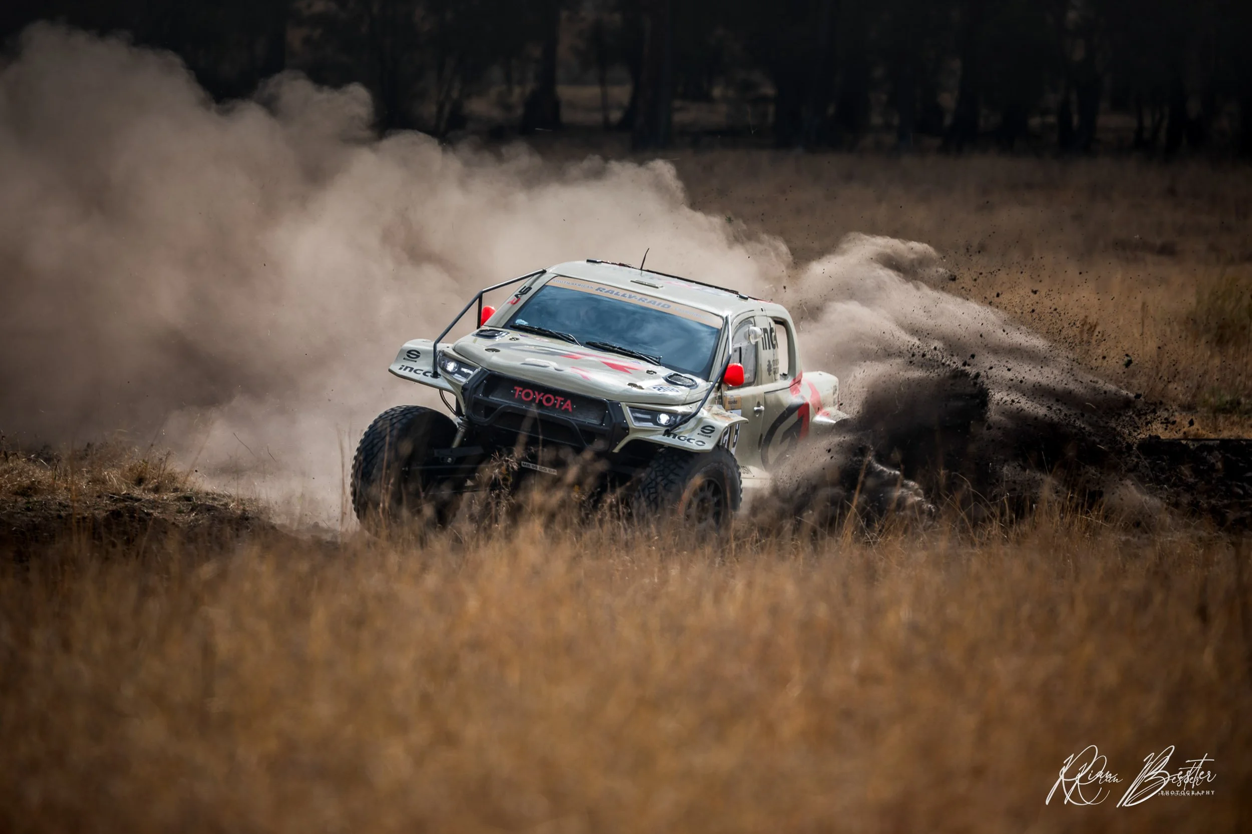 A white off-road racing truck kicking up dust as it speeds through a dirt field with trees in the background.