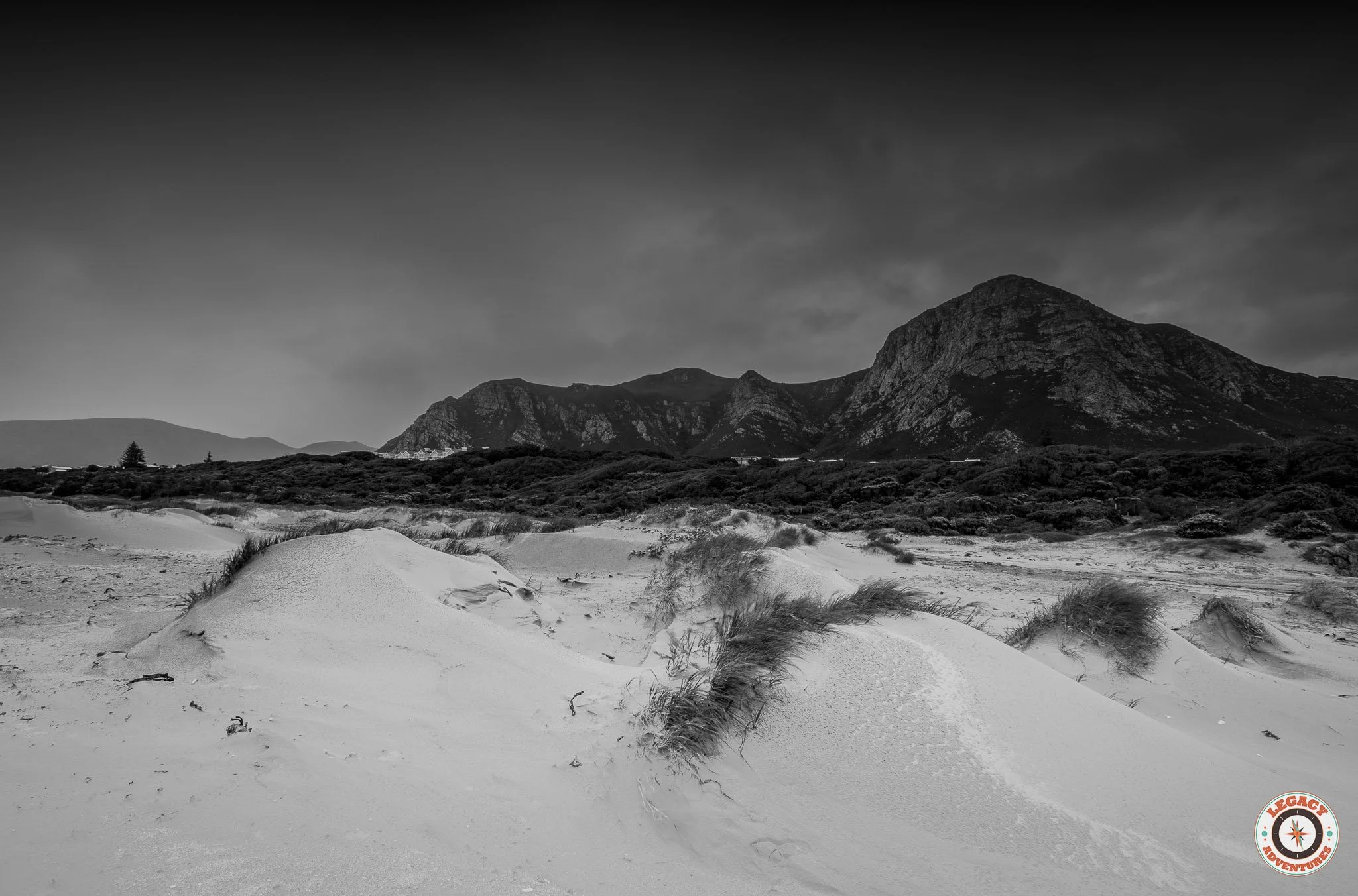 Black and white landscape of sand dunes in the foreground with mountains in the background and cloudy sky overhead, Grotto Beach, Hermanus.