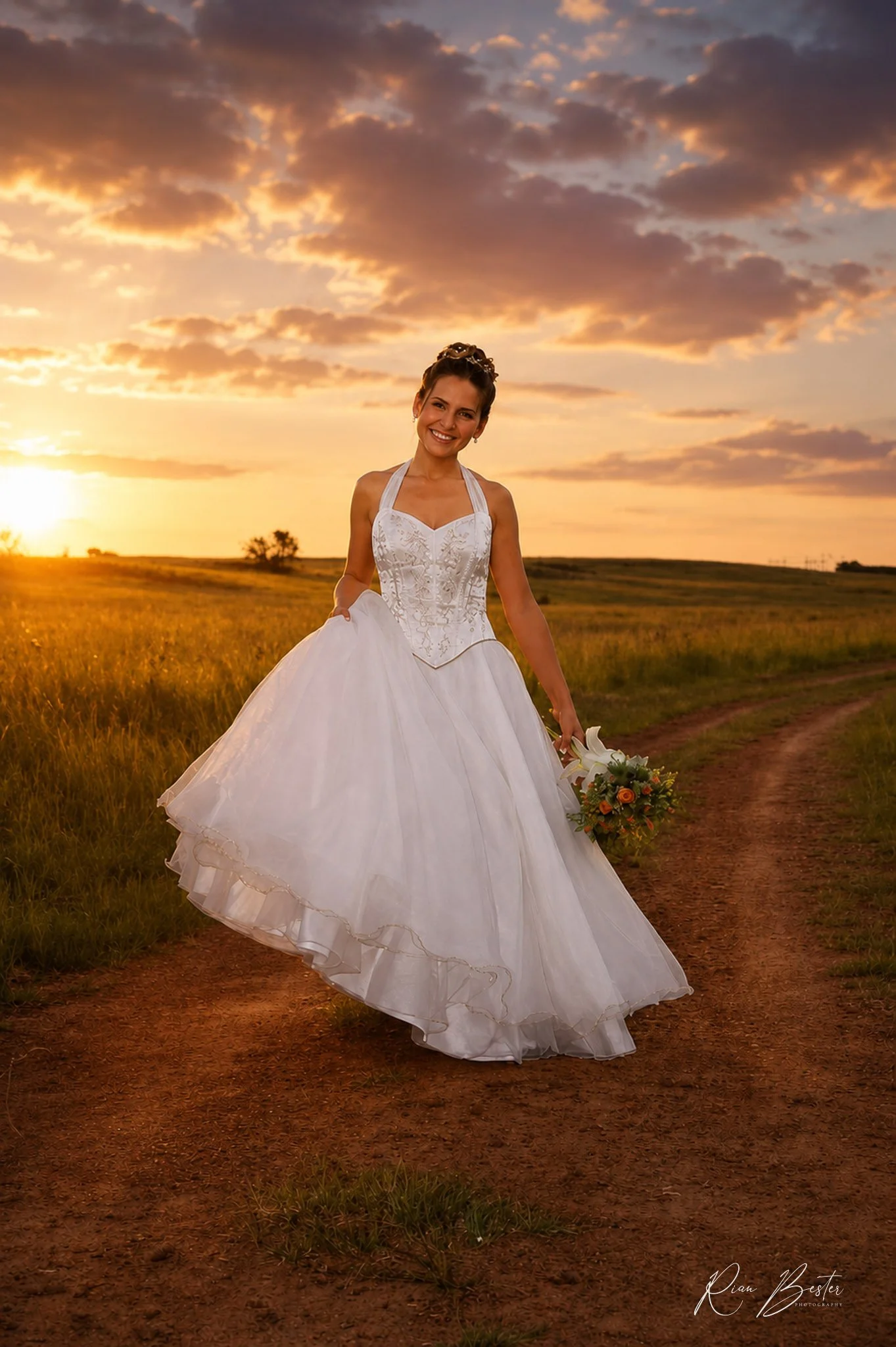 A smiling bride in a white wedding gown holding a bouquet of flowers standing on a dirt path in a grassy field at sunset, with a sky filled with clouds and the sun setting in the background.