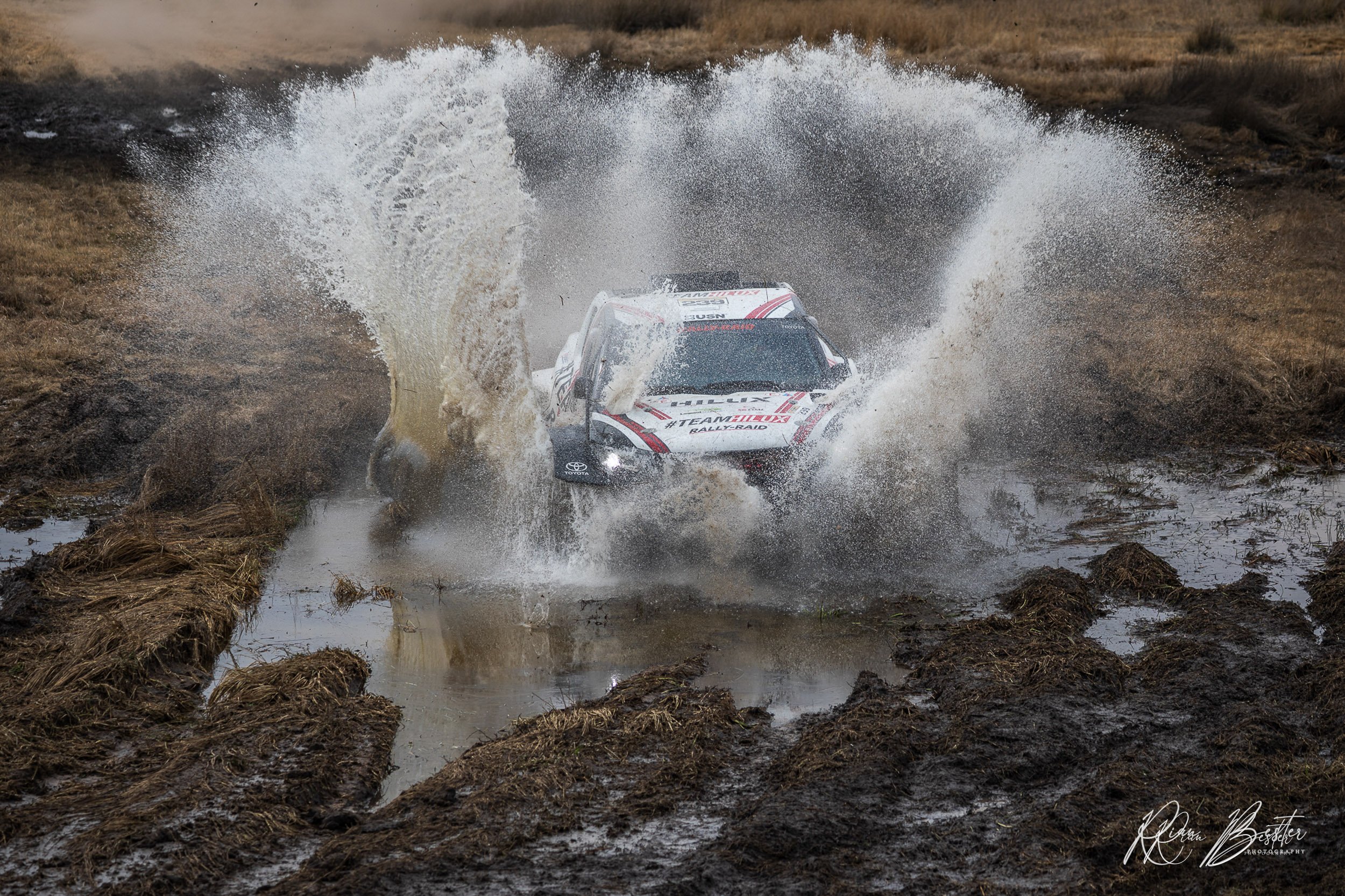 A rally car driving through a muddy water crossing, creating a large splash, in a rugged outdoor terrain.