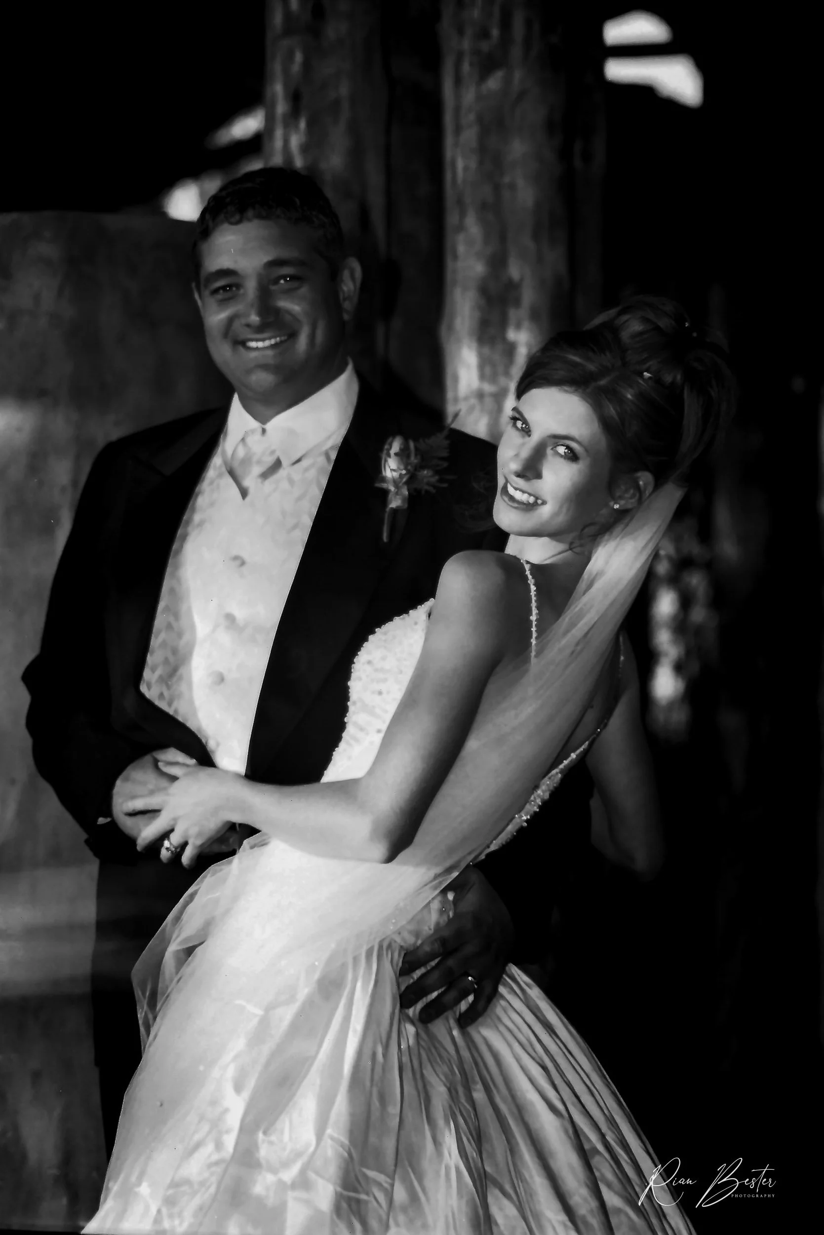 A smiling bride and groom in wedding attire, posing together in a rustic setting.