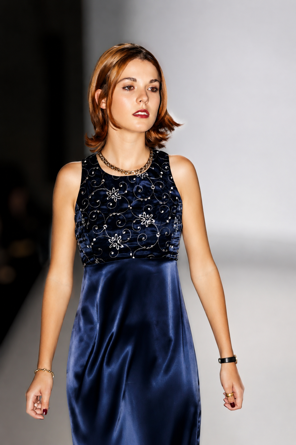 A woman with short, wavy, auburn hair wearing a sleeveless blue satin dress with floral embroidery on the top, a gold necklace, and a watch on her left wrist, walking on a runway.