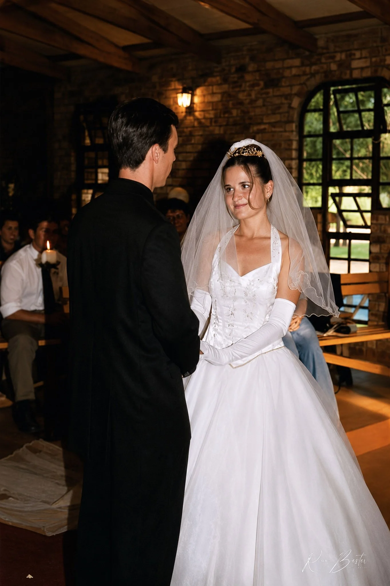 A bride and groom exchange vows in a rustic indoor setting with exposed brick walls and large windows. The bride is wearing a white wedding gown with gloves, a veil, and a tiara, while the groom is dressed in a black suit. Guests are seated in the ba