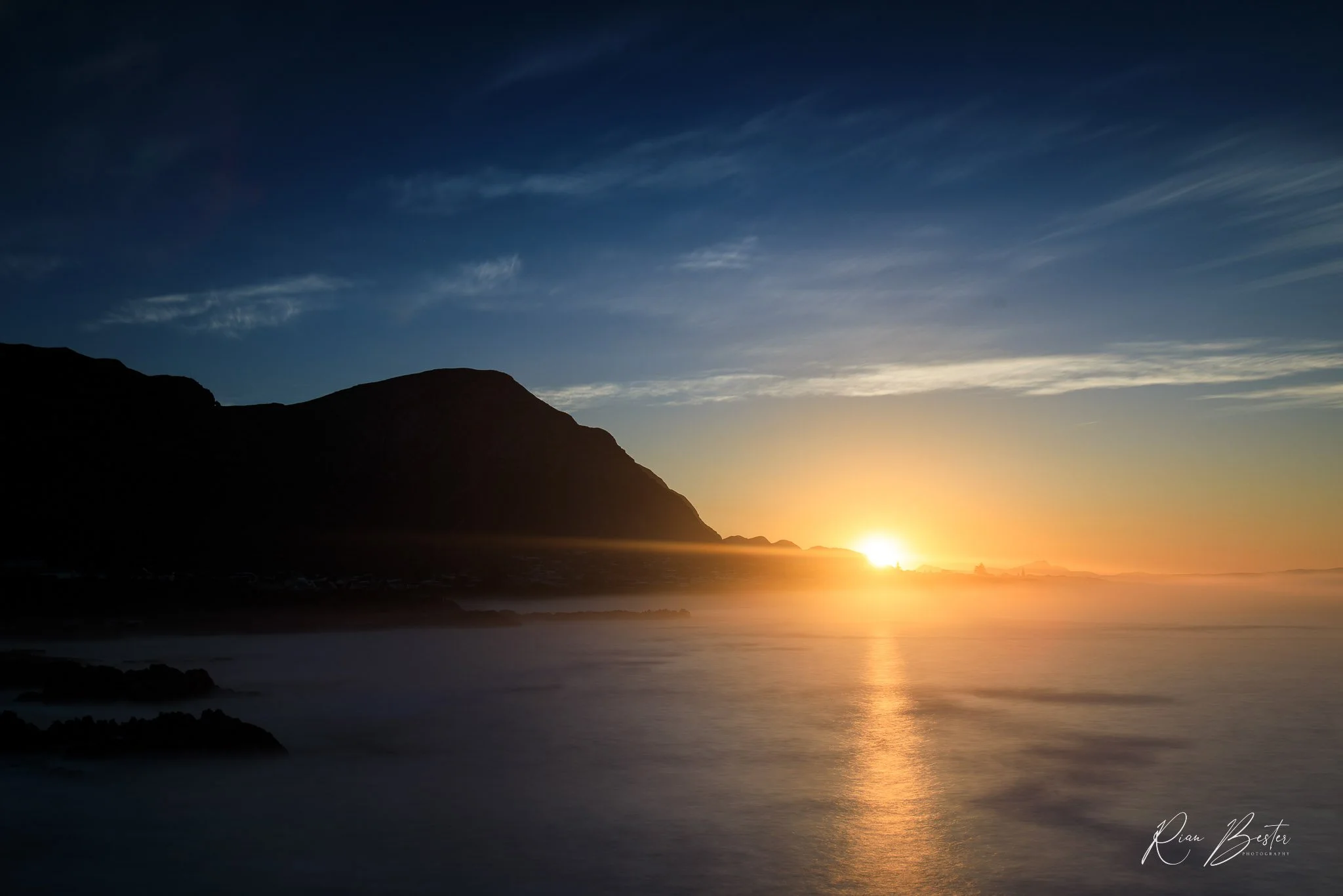 Sunset over a ocean with a mountain silhouette, blue sky with wispy clouds.