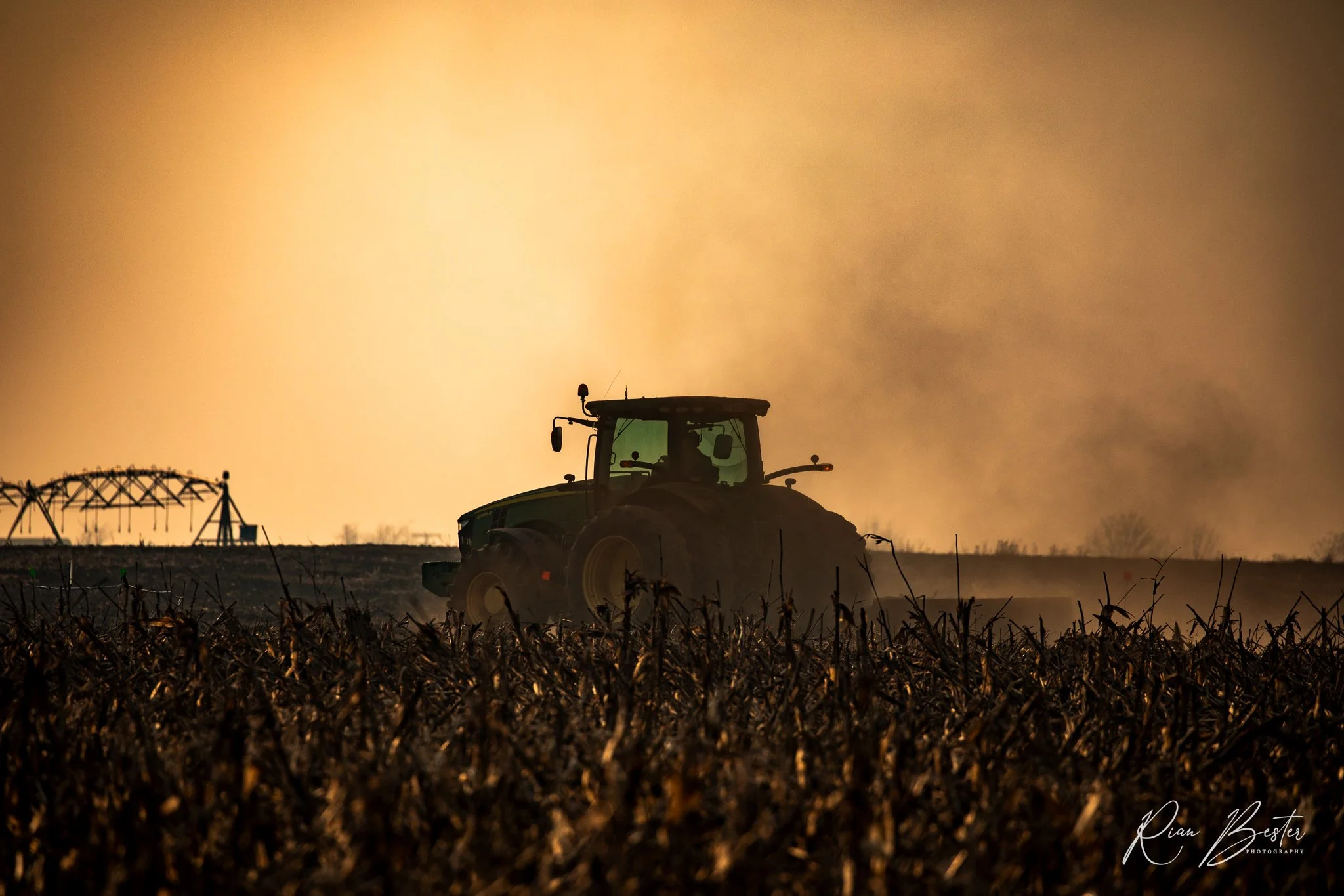 A tractor working in a field at sunset with silhouettes of farm equipment and plants in the foreground.