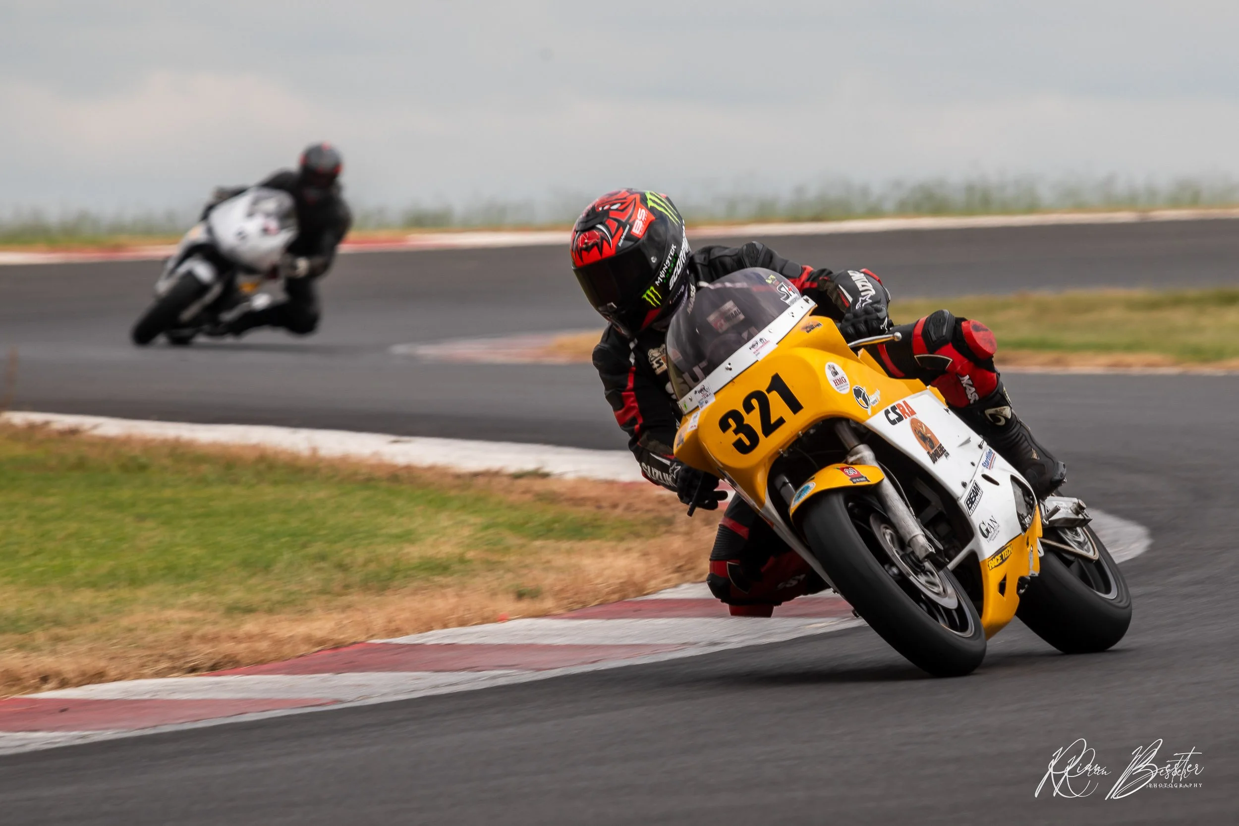 Motorcycle racers leaning into a turn on a racetrack, with the rider in the foreground on a yellow and white motorcycle numbered 321 and the rider in the background on a white motorcycle.