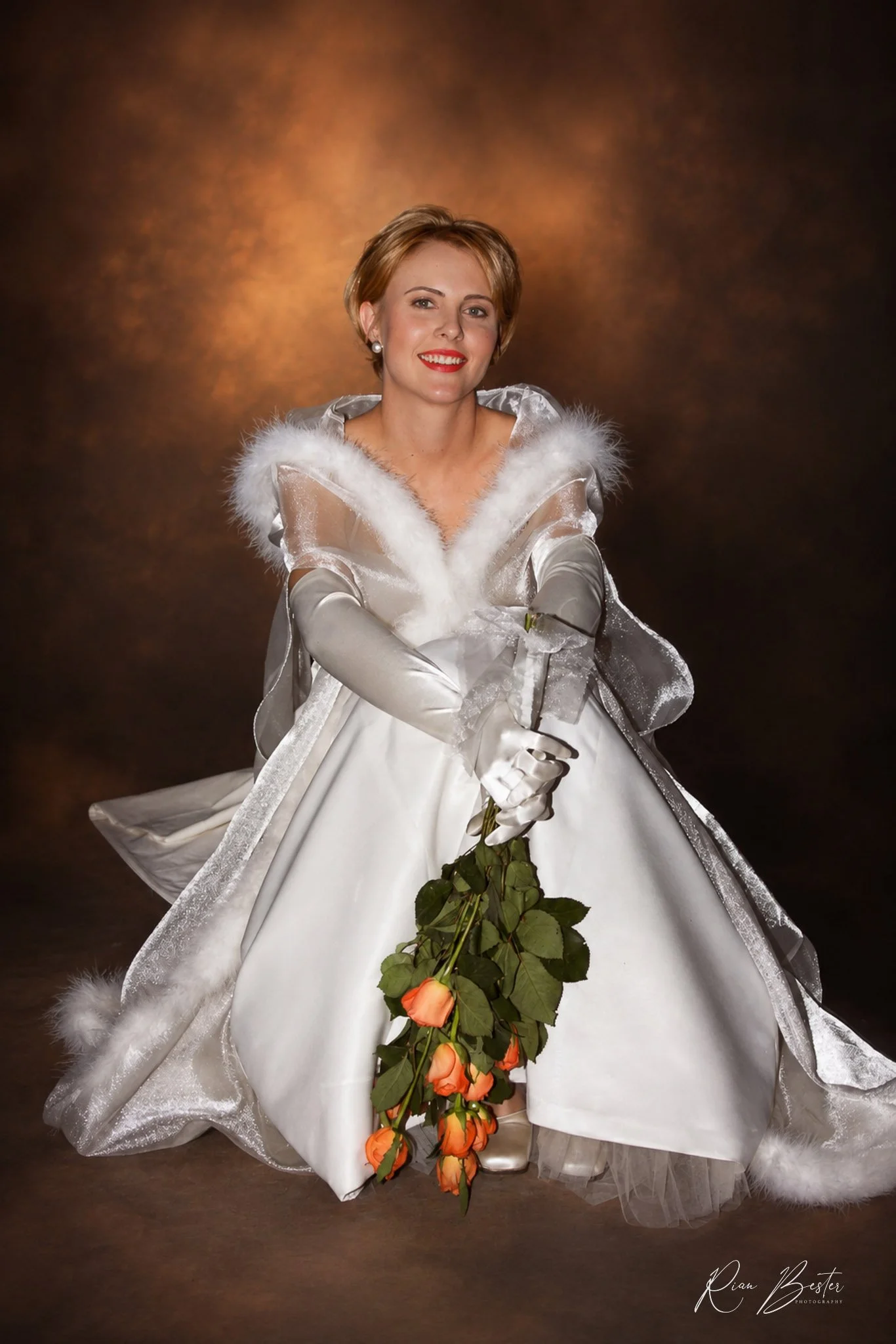 A woman in a white, satin bridal gown with fur details on the shoulders, holding a bouquet of orange roses, smiling in front of a brown mottled background.