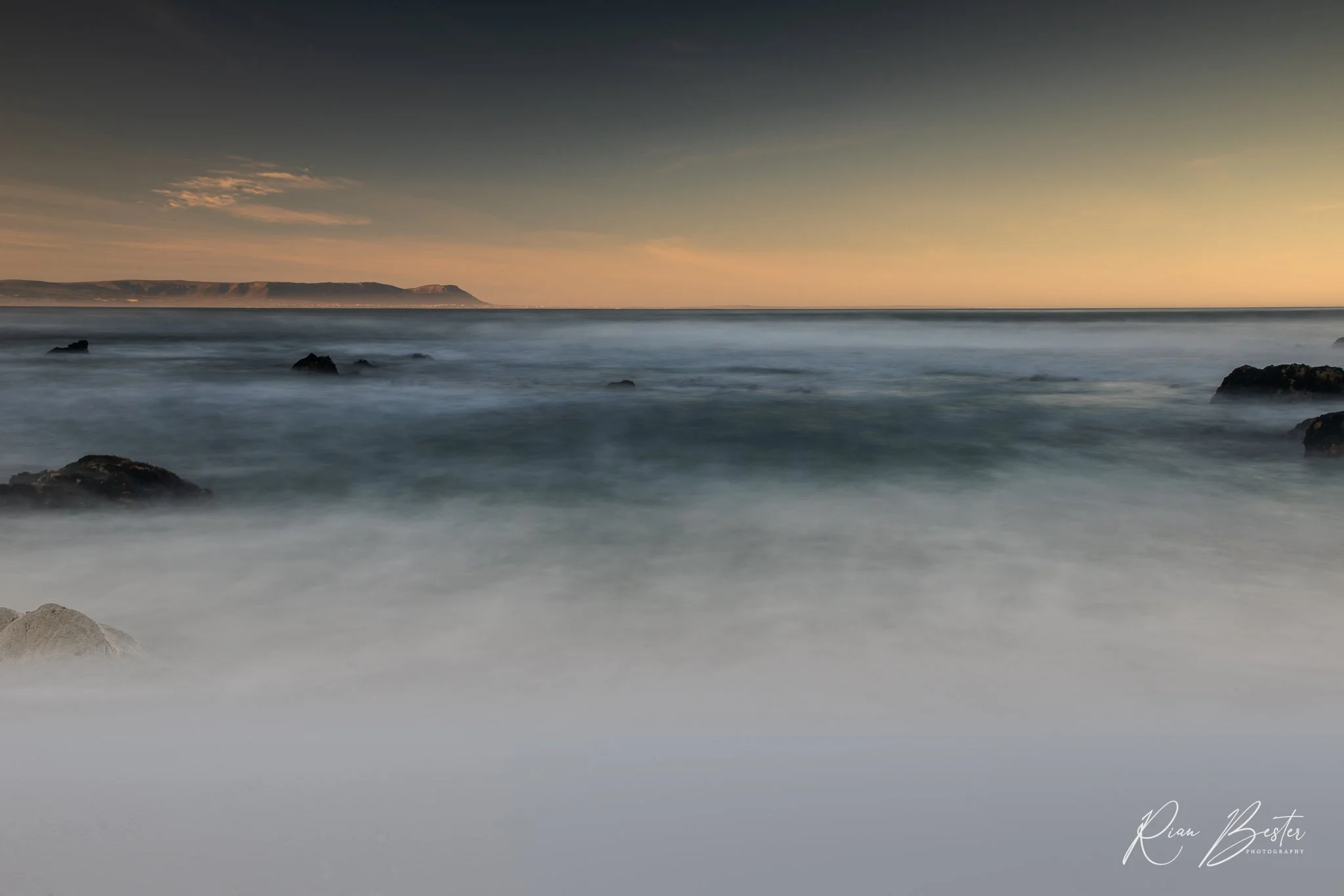 Long exposure photograph of the ocean at sunset with calm waves and distant mountains on the horizon. Rock formations are visible in the water, and the sky has a gradient of warm and cool tones.