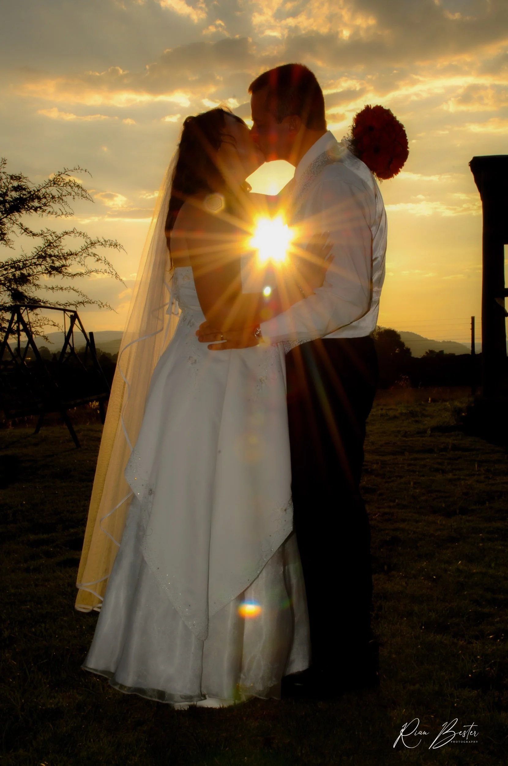 A bride and groom sharing a kiss during sunset, with the sun shining between them, on their wedding day.