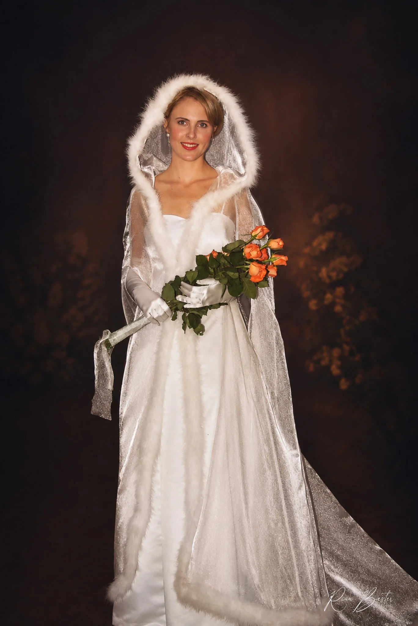 A woman dressed in a white wedding gown with a hooded fur-lined cape, holding a bouquet of orange roses, smiles against a dark background.