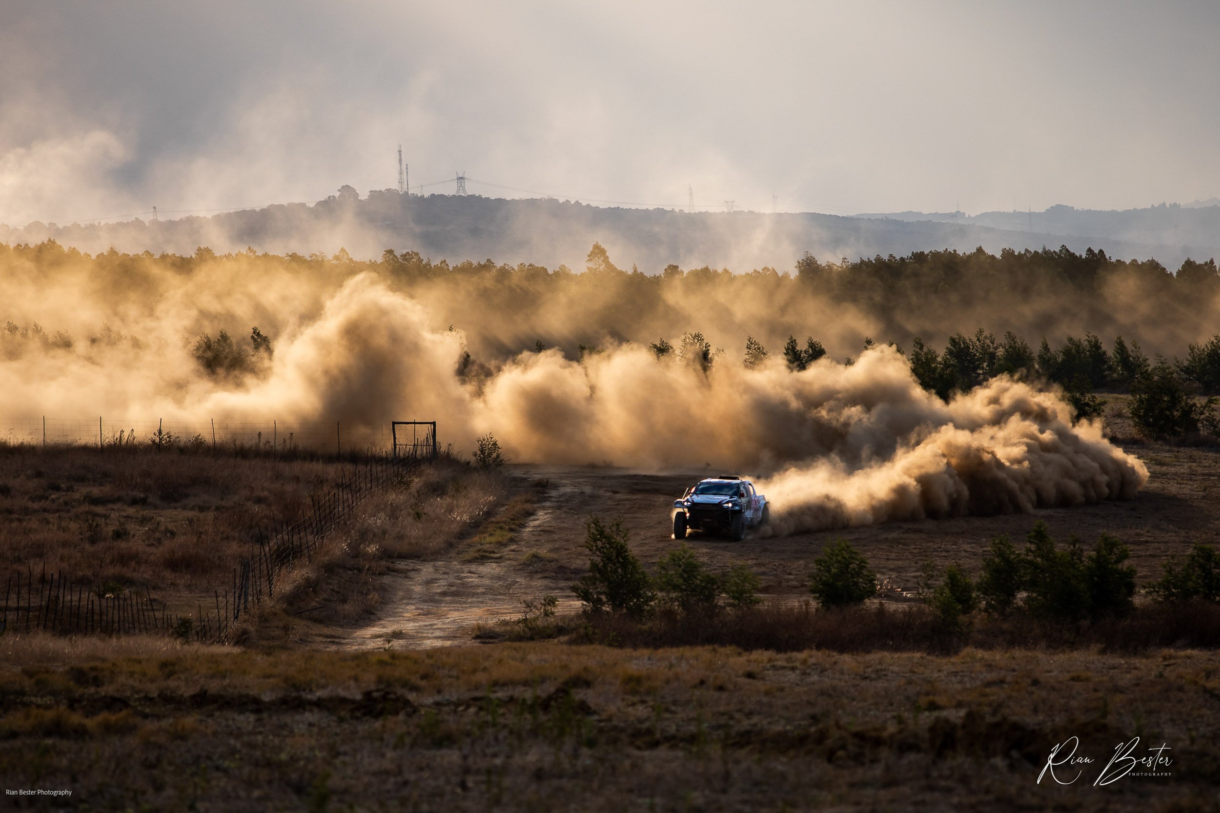 A black off-road truck speeding on a dirt path, kicking up a large cloud of dust amidst a dry field and sparse trees, with a distant tree line and hills in the background.
