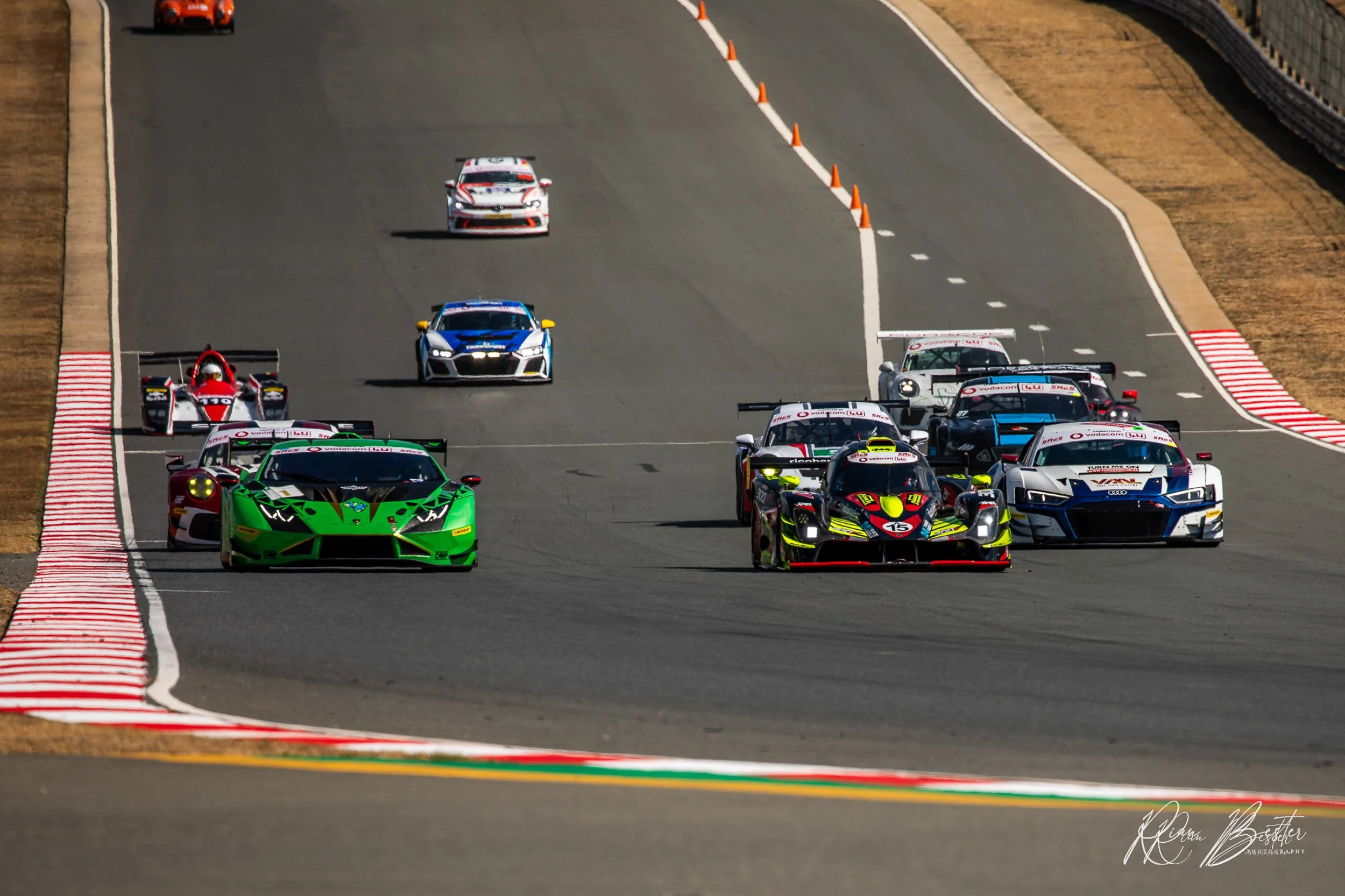 Multiple race cars competing on a racetrack, including a green Lamborghini, a black and red race car, a white Audi, and other cars in the background, with orange cones and track markings.