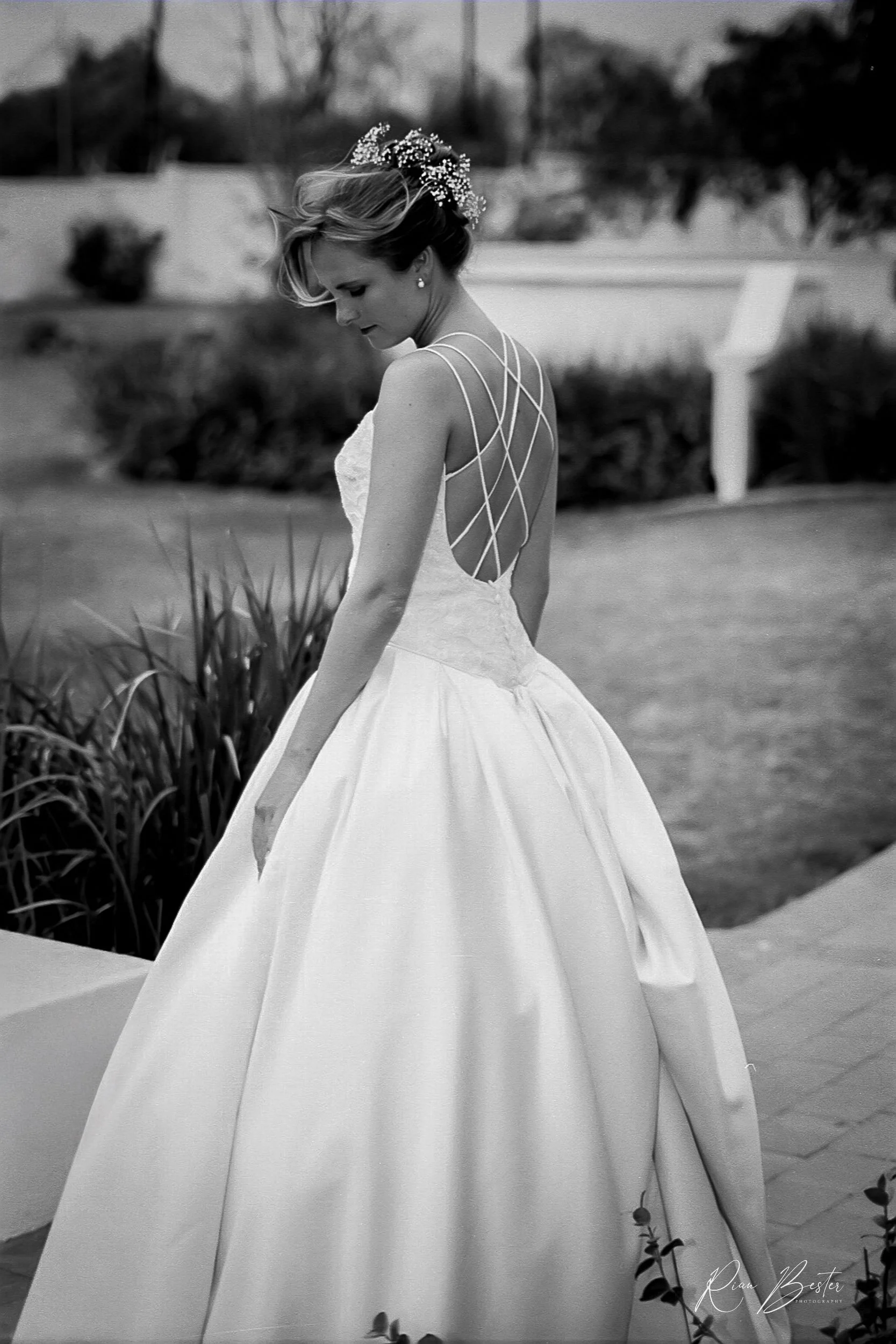 A woman wearing a wedding dress with a lace bodice and a full skirt, standing outdoors with a floral hair accessory and looking down.