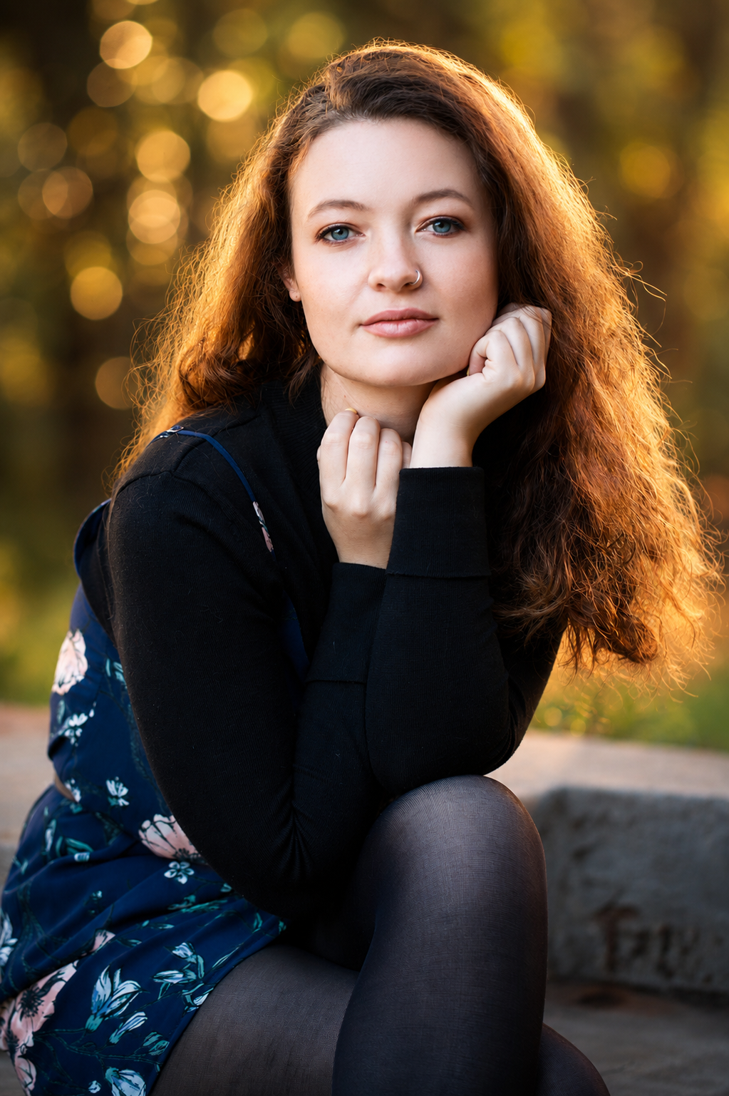 A young woman with curly red hair and blue eyes sitting outdoors at sunset, resting her chin on her hand, with warm golden light and blurred autumn trees in the background.