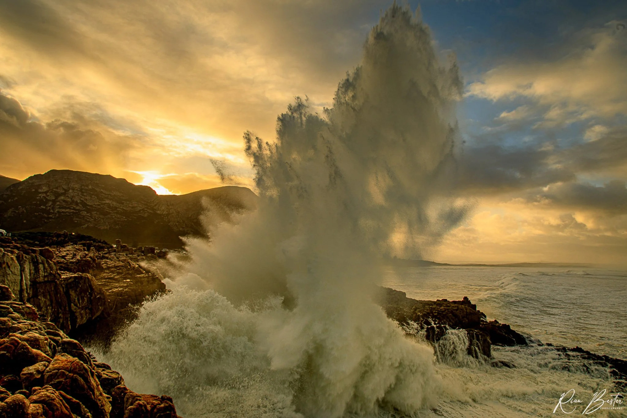 A sunset over the ocean with waves crashing against rocky cliffs, creating large spray, and a mountainous landscape in the background.