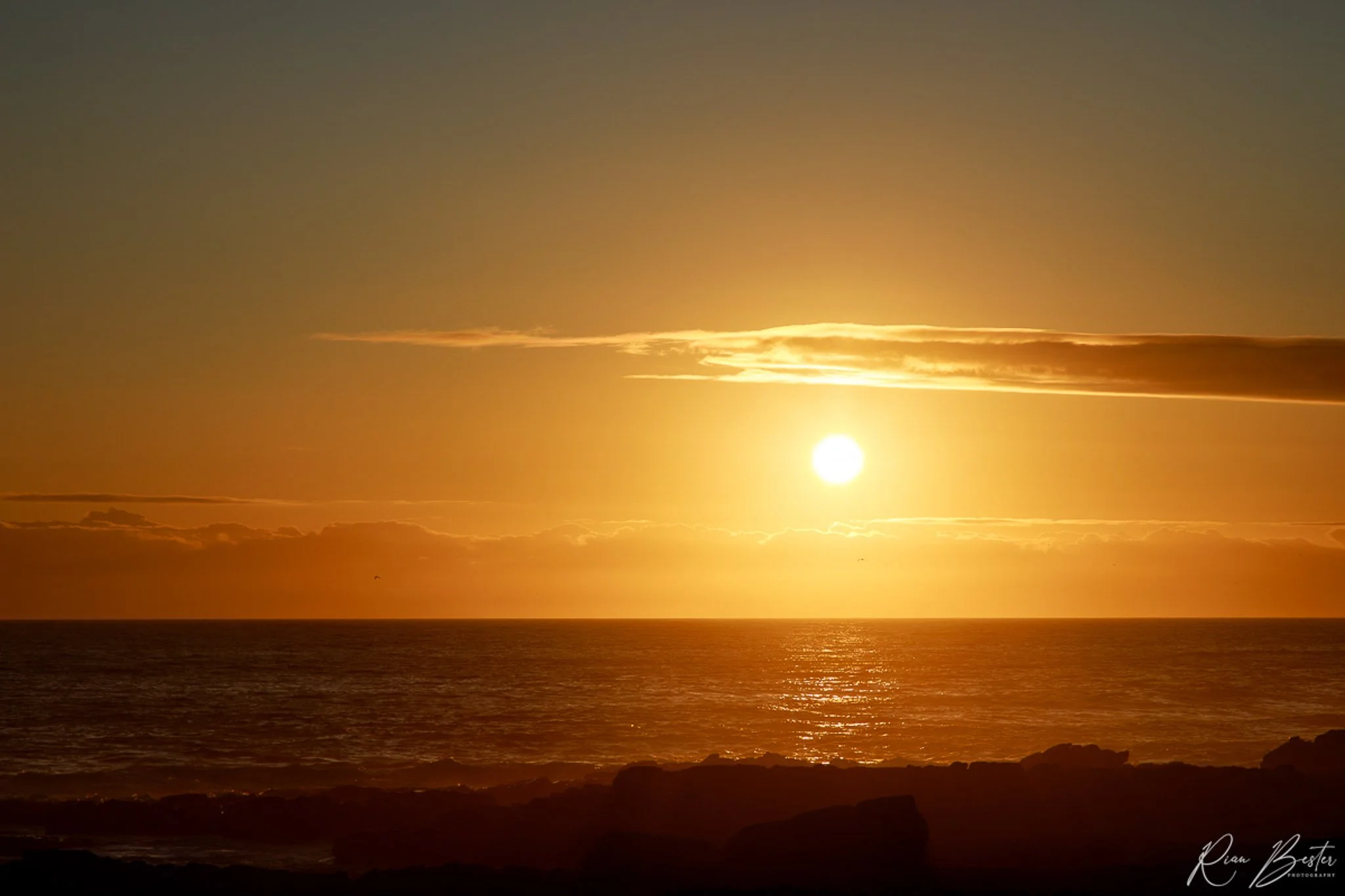 Sunset over the ocean with clouds in the sky and rocks on the shoreline in silhouette.