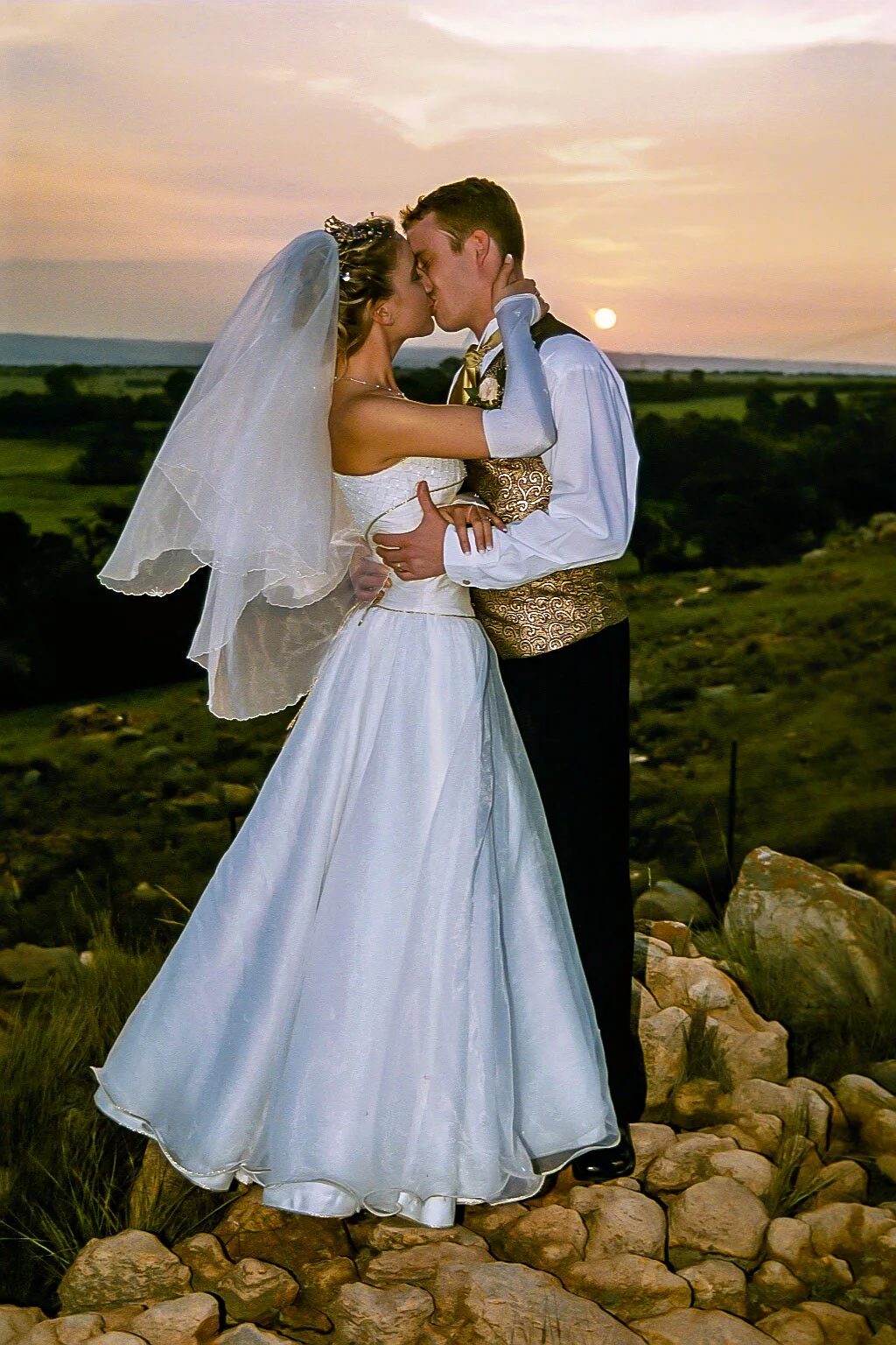 A bride and groom sharing a kiss at sunset in a rural landscape, with rocks in the foreground.