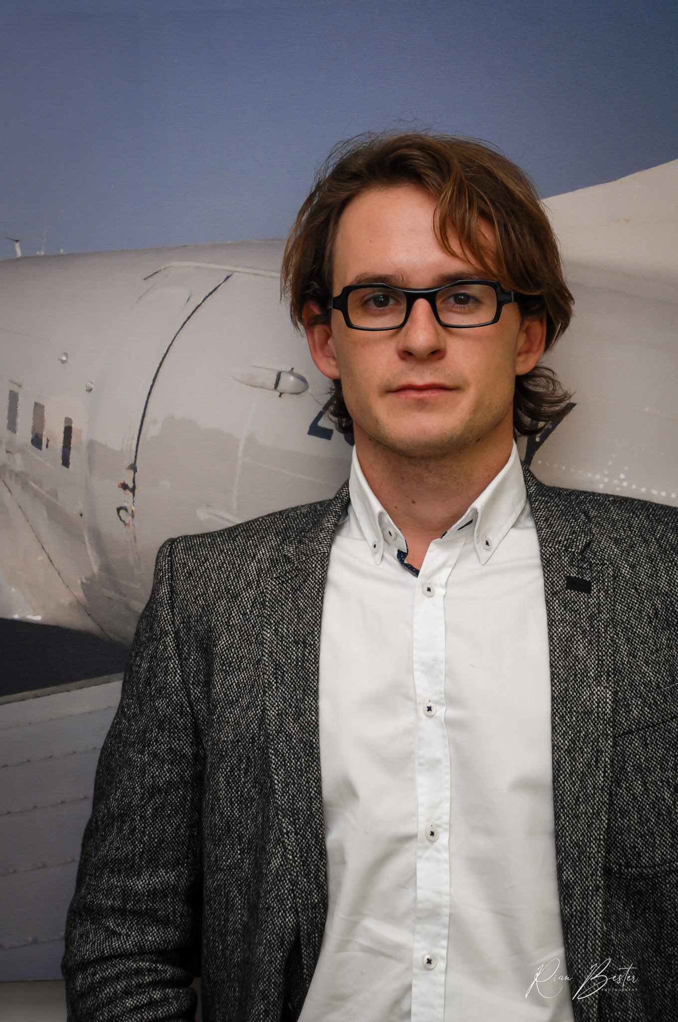 A young man with glasses and brown wavy hair standing in front of an airplane fuselage with a blue sky above.