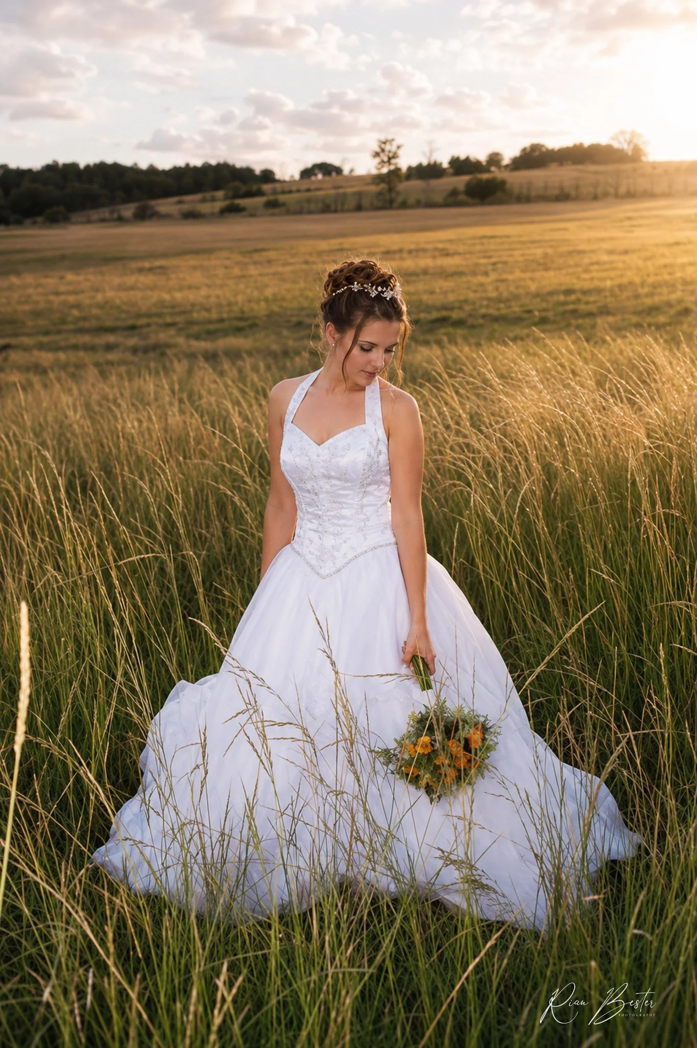 A woman in a white wedding gown stands in a field of tall grass during sunset, holding a bouquet of flowers.