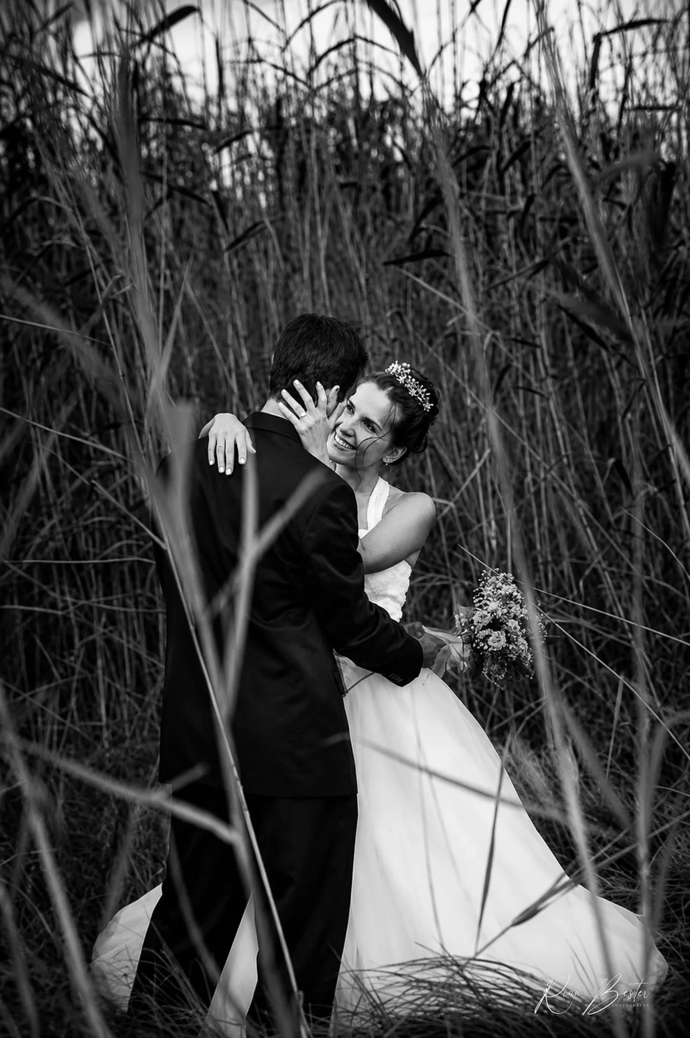 Black and white photo of a couple dressed in wedding attire embracing in tall grass or reeds outdoors, with the woman smiling at the man.