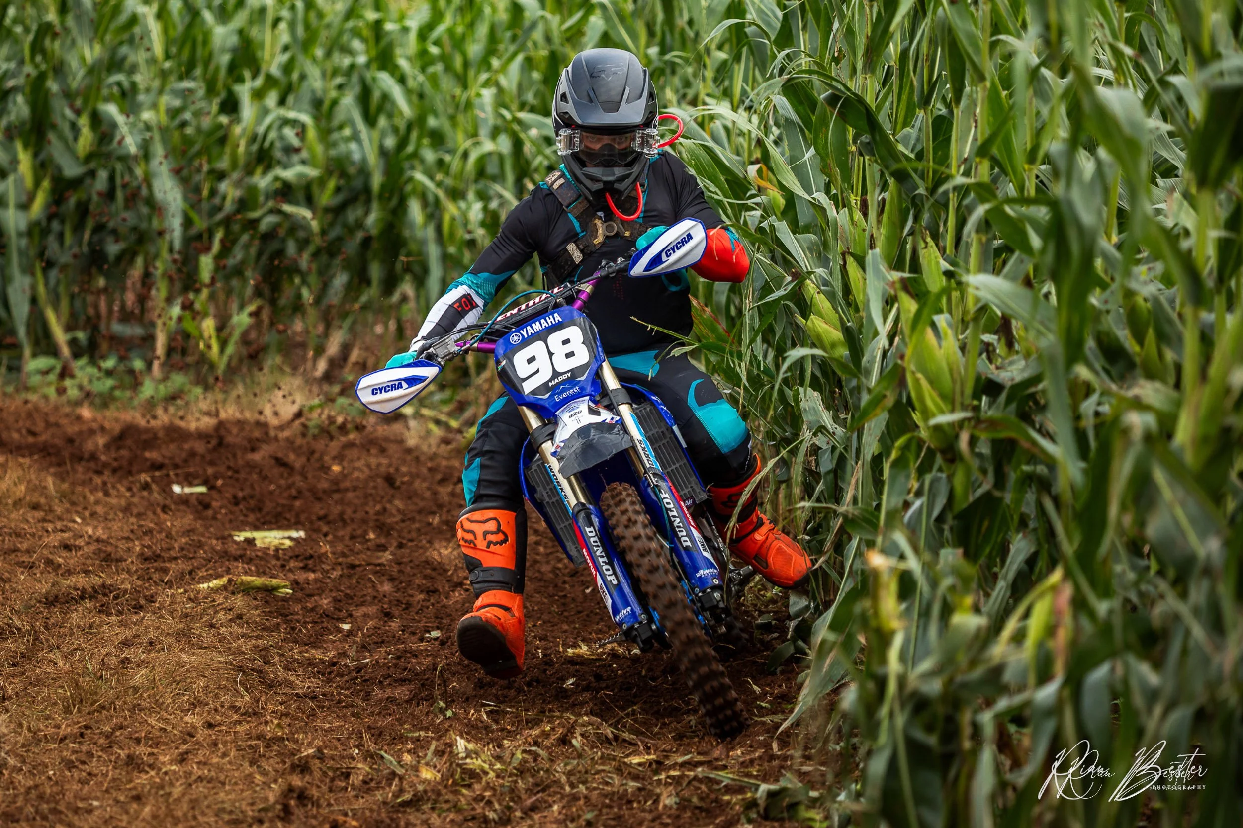 A motocross rider wearing a black helmet, black and blue racing gear, and bright orange boots navigates a dirt path through a cornfield on a blue Yamaha dirt bike with the number 98. The rider is leaning into a turn on the trail.
