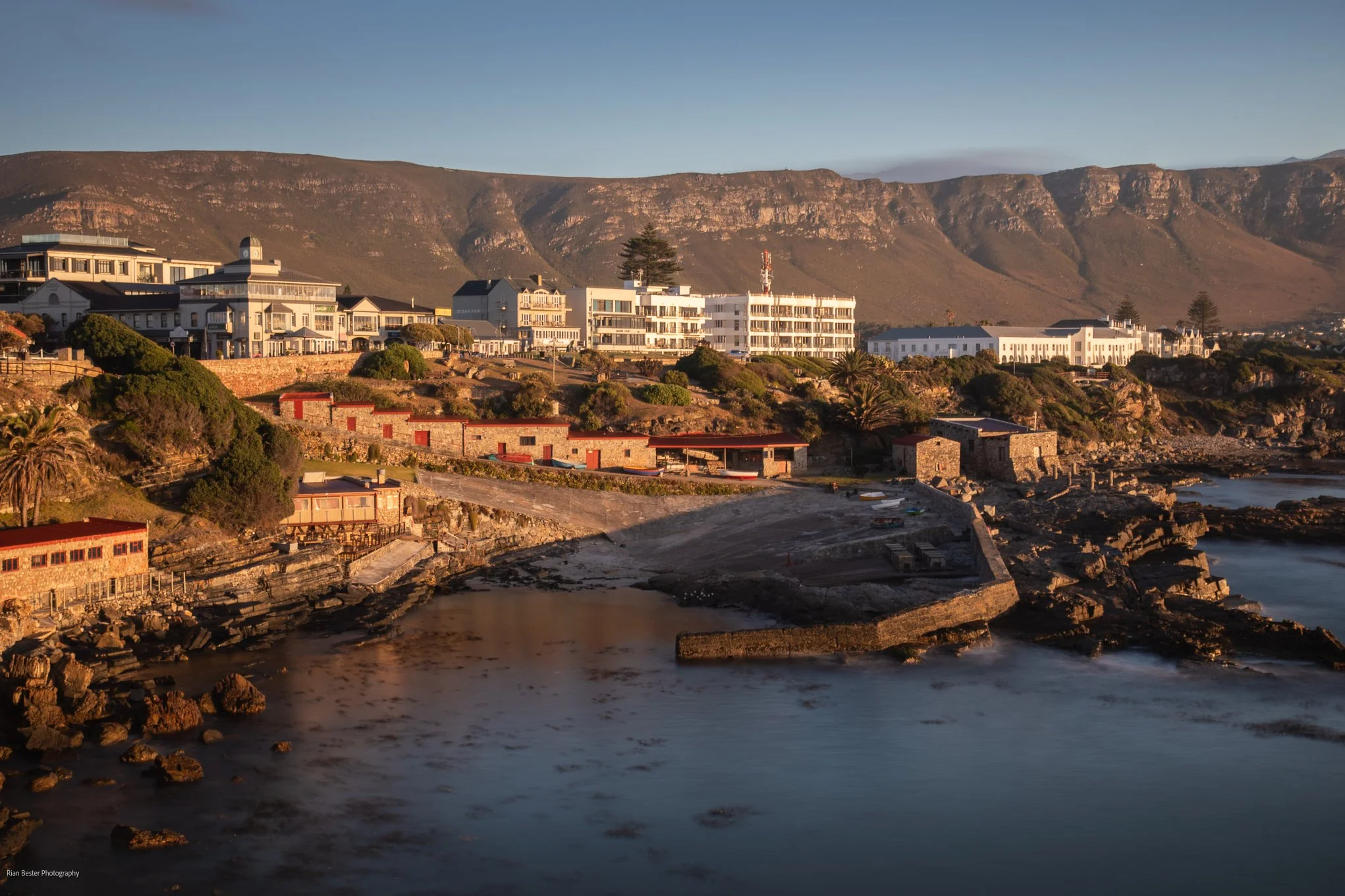 Coastal scene with white buildings along the shoreline, rocky beach, and mountains in the background during sunset.