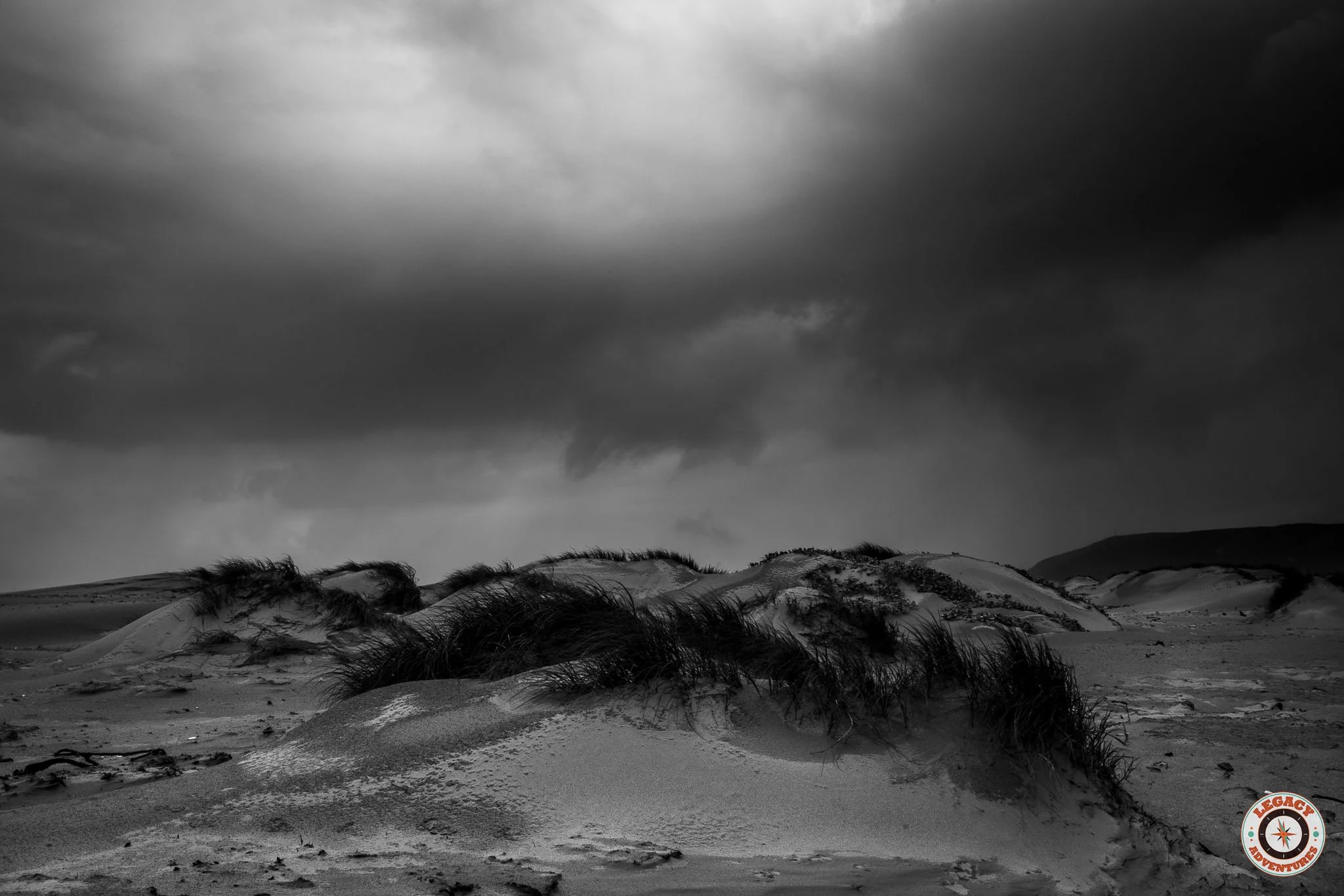 Black and white image of sand dunes with sparse grass under a stormy, cloudy sky.