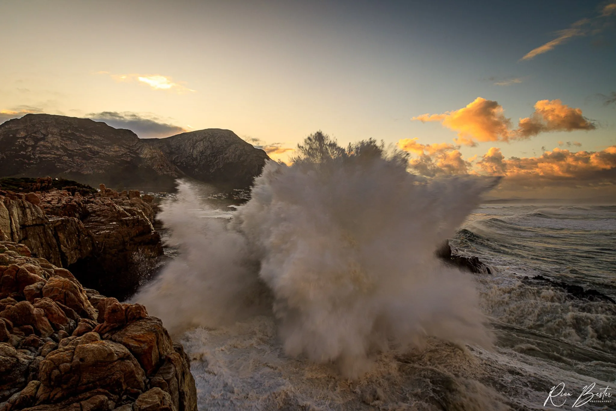 Rough ocean waves crashing against rocky shoreline during sunset with mountains in the background and a partly cloudy sky.
