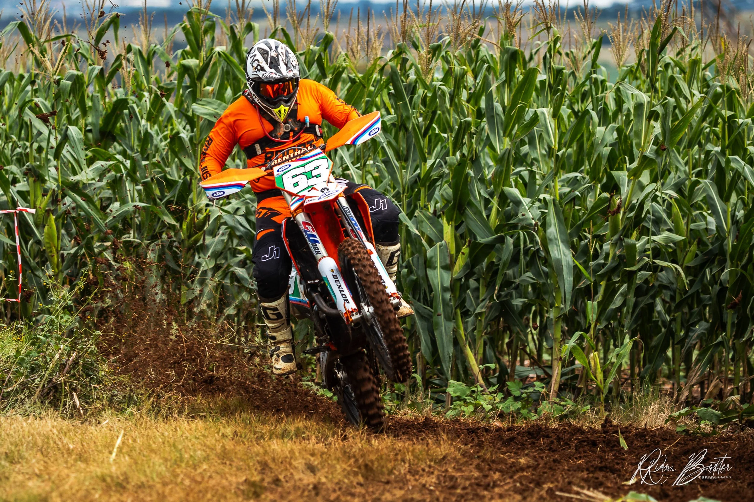 Motocross rider in orange gear and helmet riding a dirt bike through a cornfield.