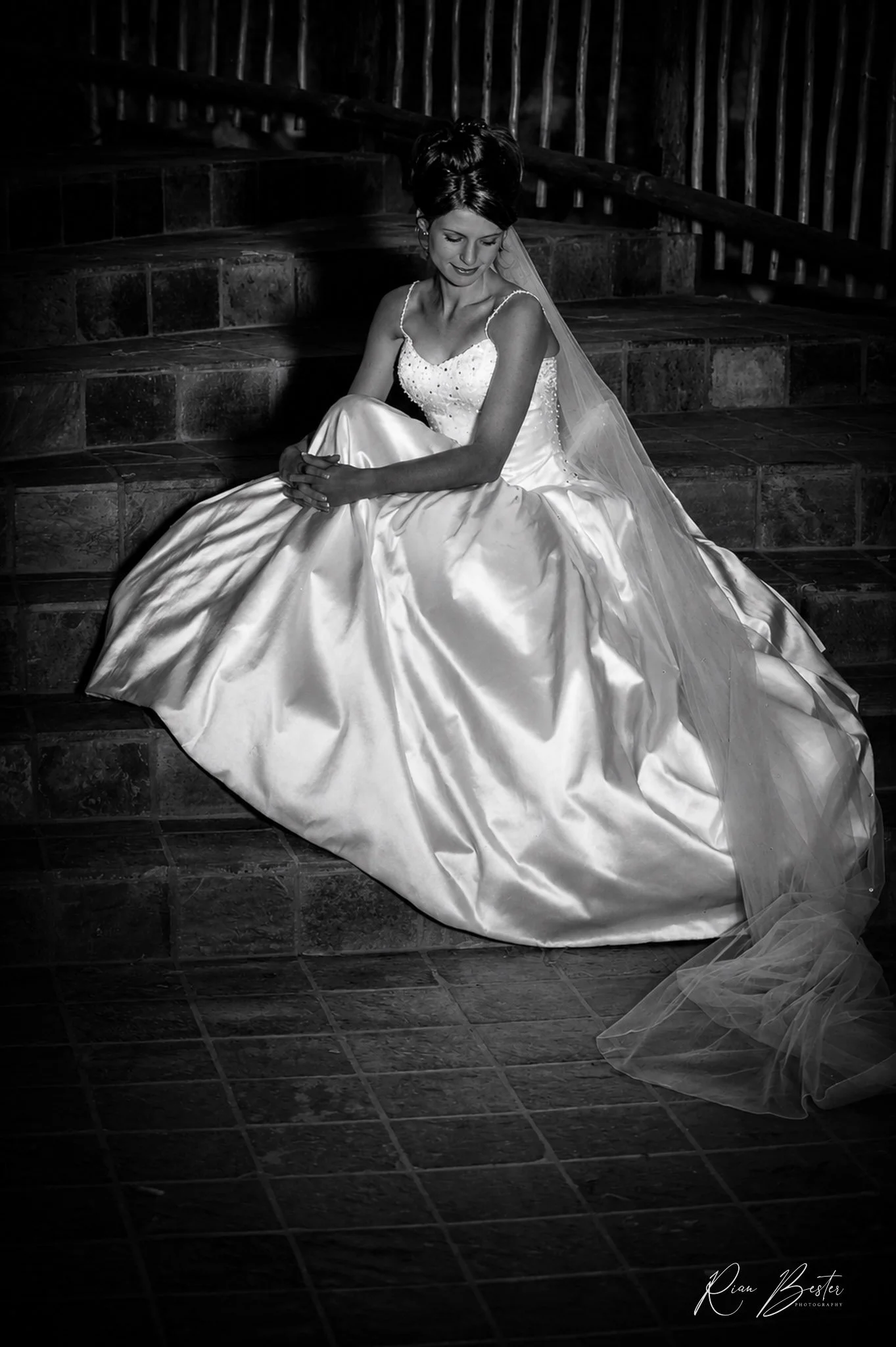 Black and white photo of a woman in a wedding dress sitting on stairs with a wooden railing in the background, smiling and looking down.