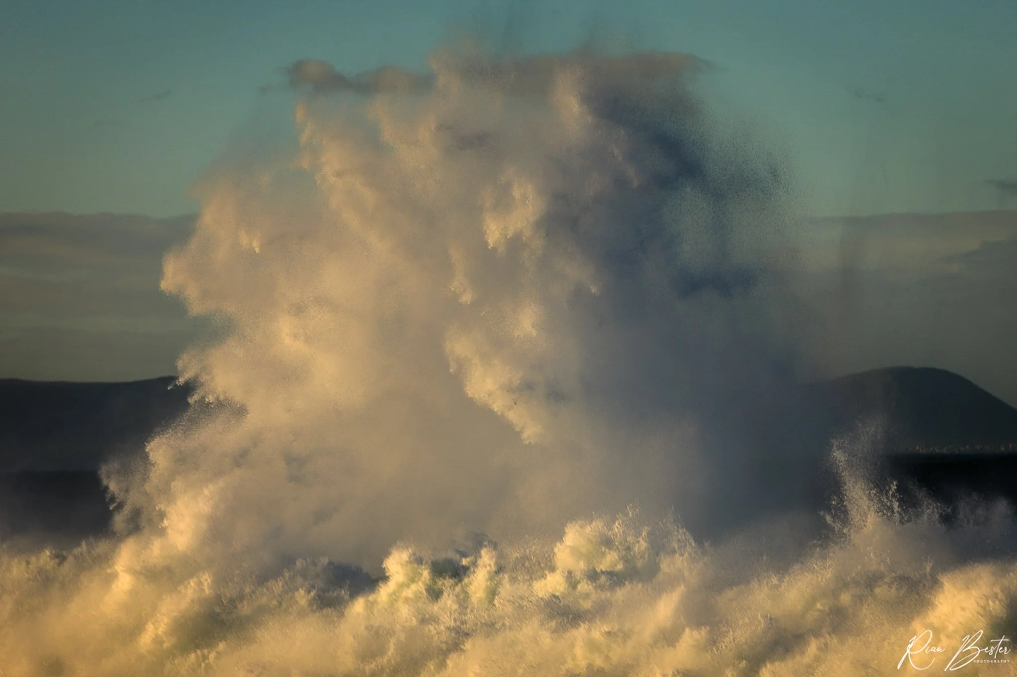 Ocean waves crashing with large spray and foam against a backdrop of dark mountain silhouettes.
