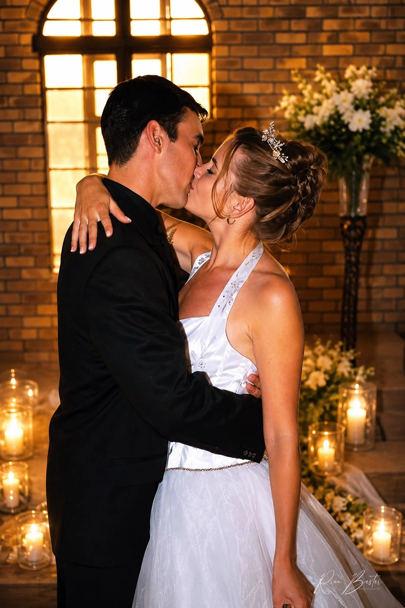 A bride and groom share a kiss during their wedding ceremony. The bride is wearing a white wedding dress with lace details and a tiara, and the groom is dressed in a black suit. The background features a lit candle arrangement and a brick wall with a