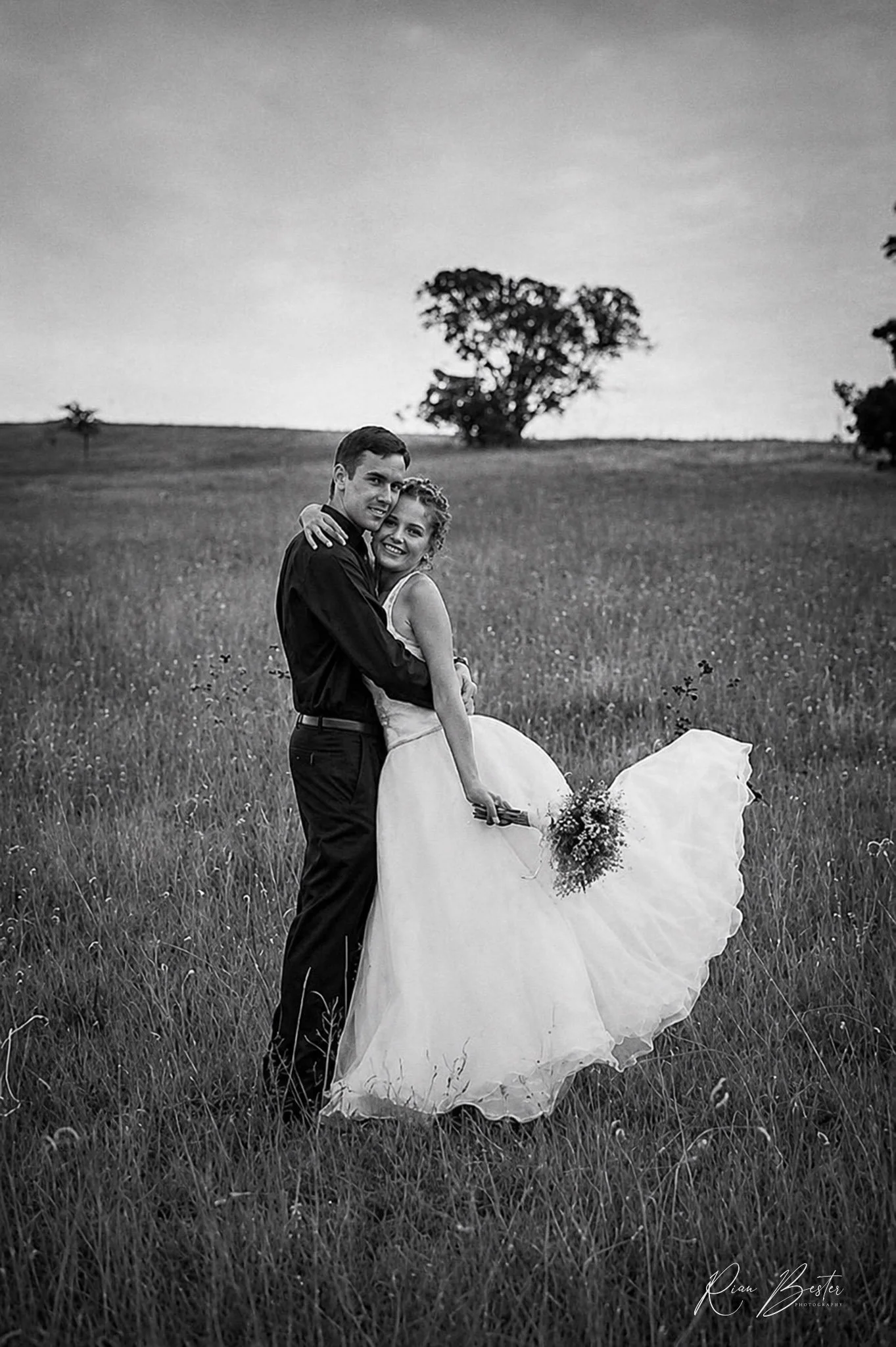 A black and white photo of a couple on their wedding day standing in a grassy field. The groom is holding the bride, who is holding a bouquet of flowers, and they are smiling at the camera with a large tree and a cloudy sky in the background.