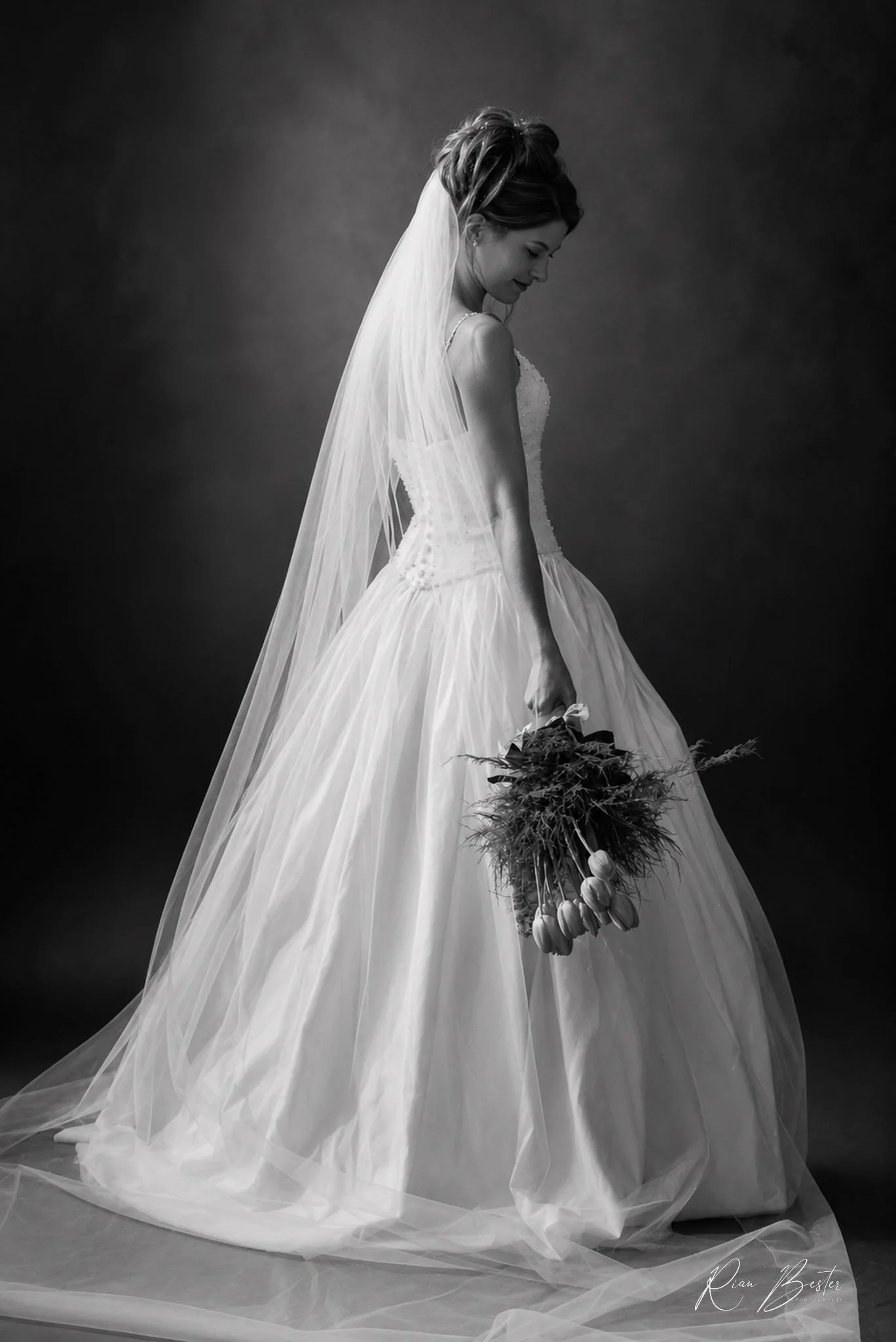 Black and white photo of a bride in a wedding dress holding a bouquet of flowers while looking down. The bride has her hair styled up and wears a veil. The dress has a fitted bodice and a full, flowing skirt with a tulle overlay.