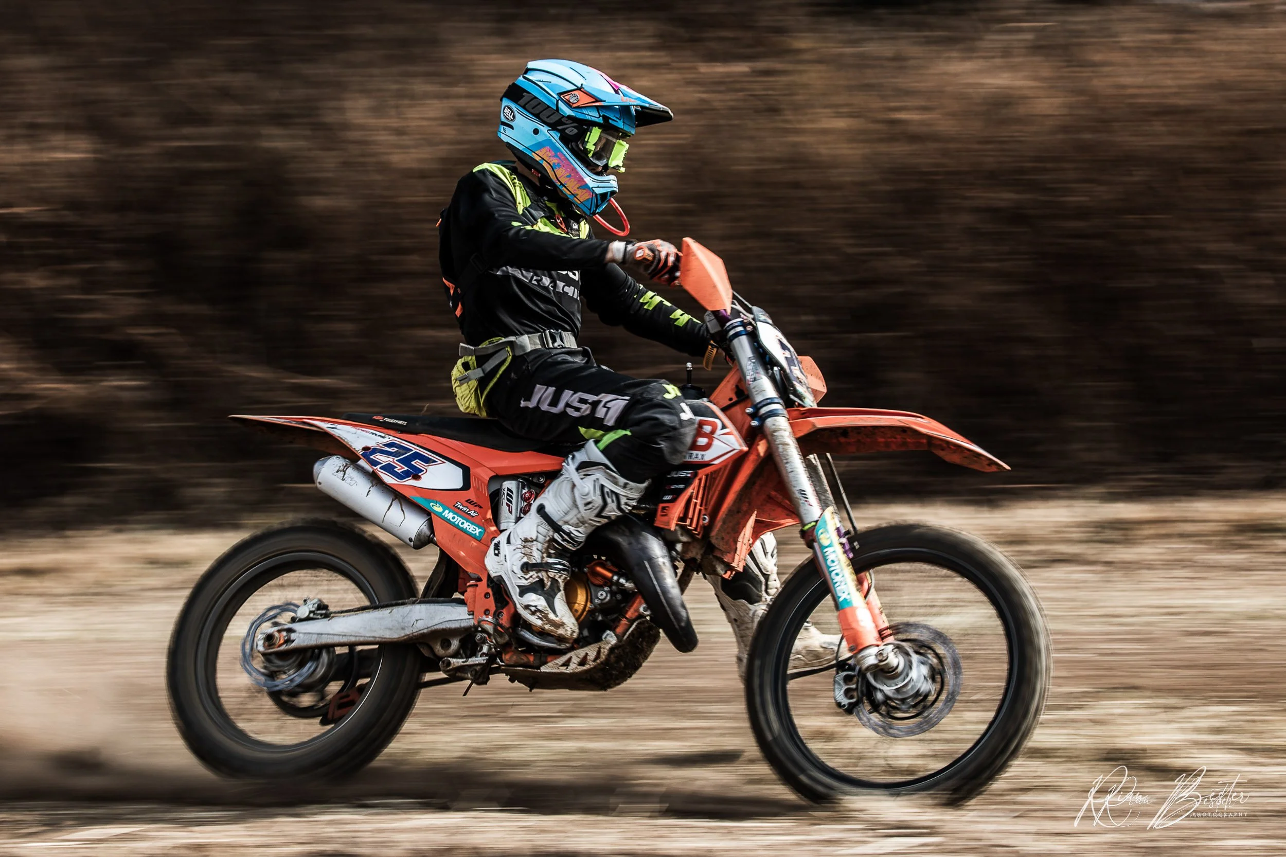 A person riding a dirt bike on a track, wearing a blue helmet and black riding gear with green accents, with a blurred background indicating motion.