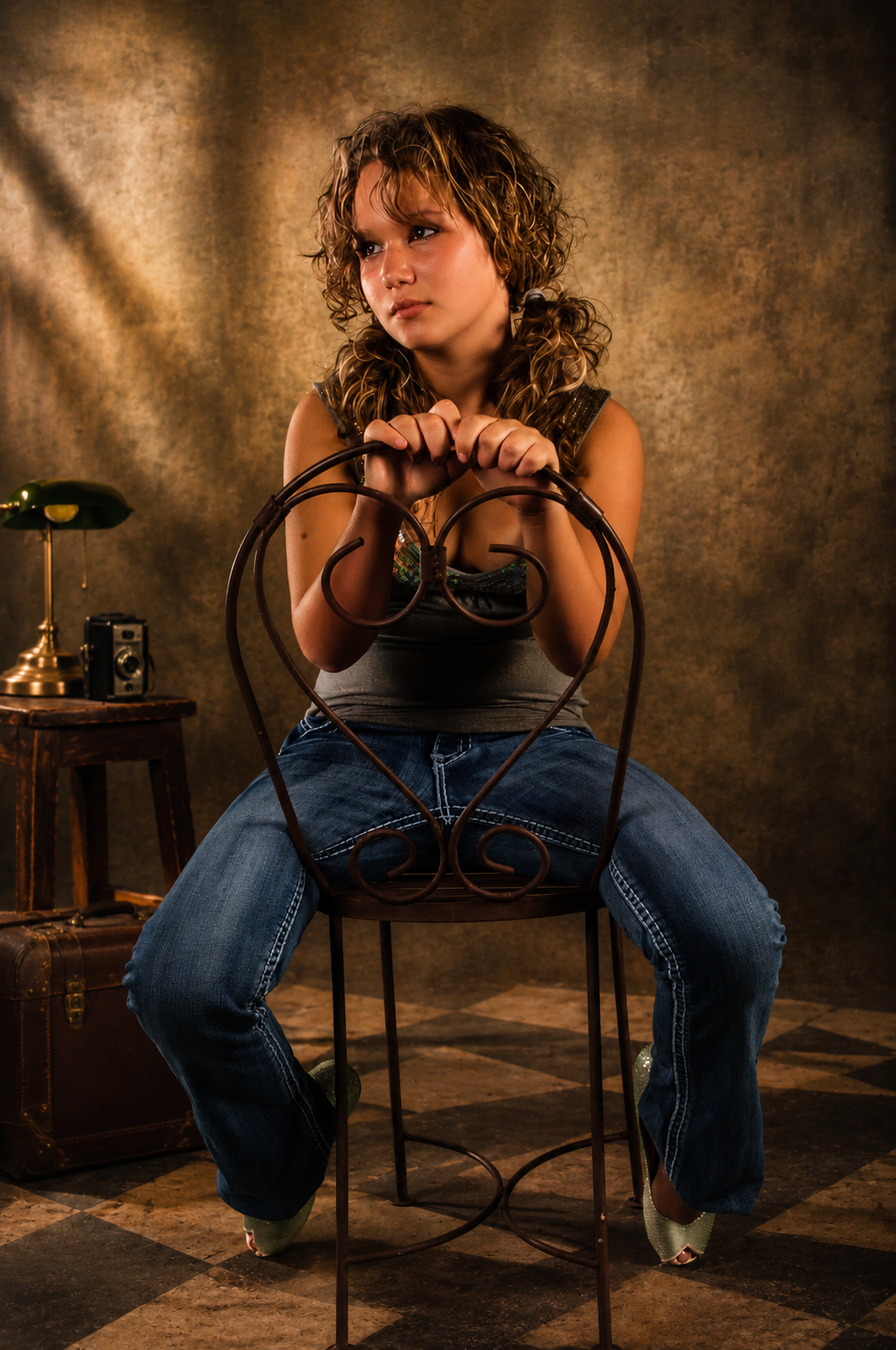 A young woman with curly hair, wearing a tank top and jeans, sitting on a metal chair, holding onto the backrest with a thoughtful expression. There is a vintage table with a lamp and a camera in the background. The setting appears to be a studio wit