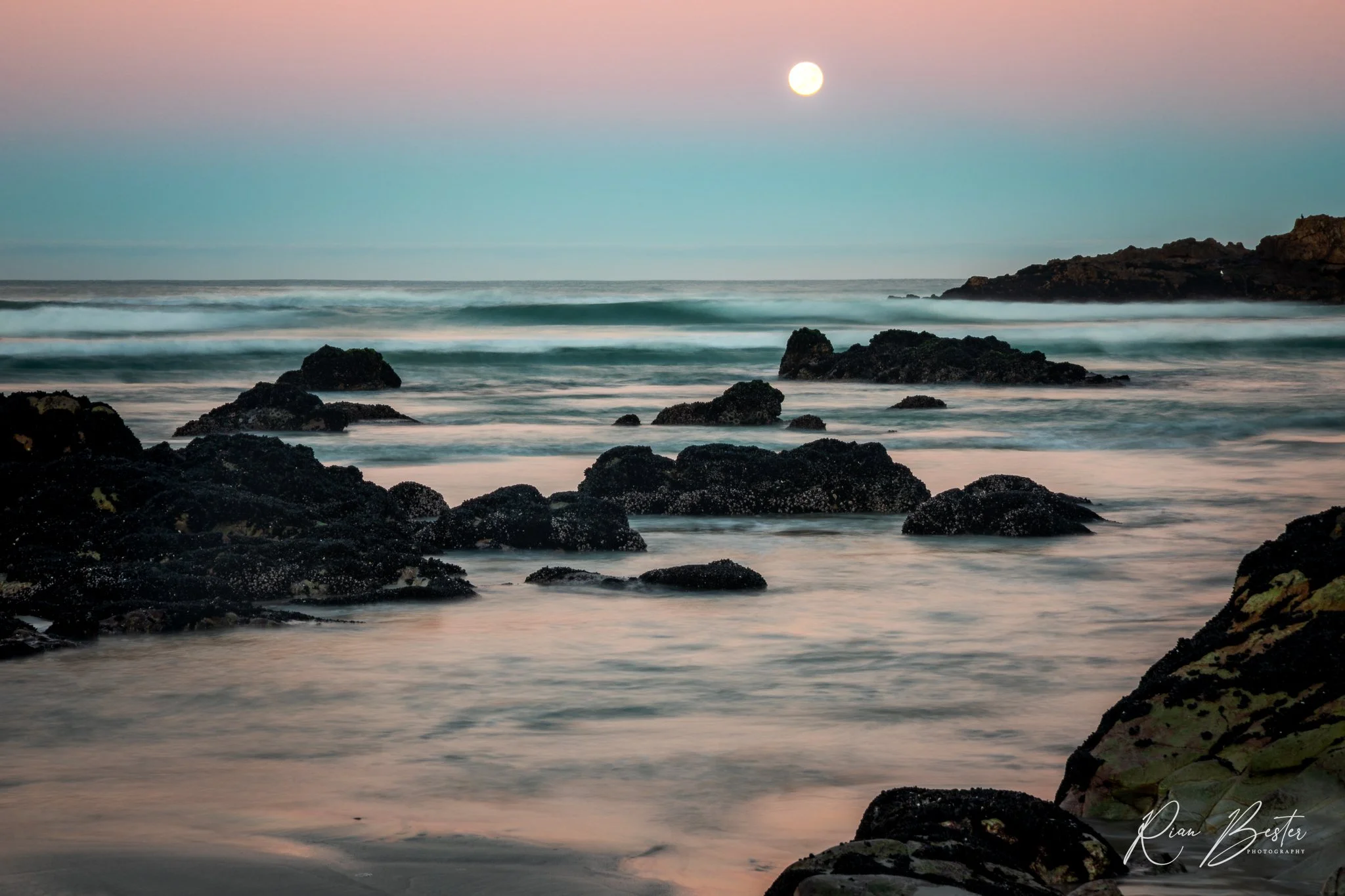 A serene ocean scene at dusk with a full moon in the sky, gentle waves hitting dark rocks along the shoreline, and a pastel-colored sky transitioning from pink to blue.
