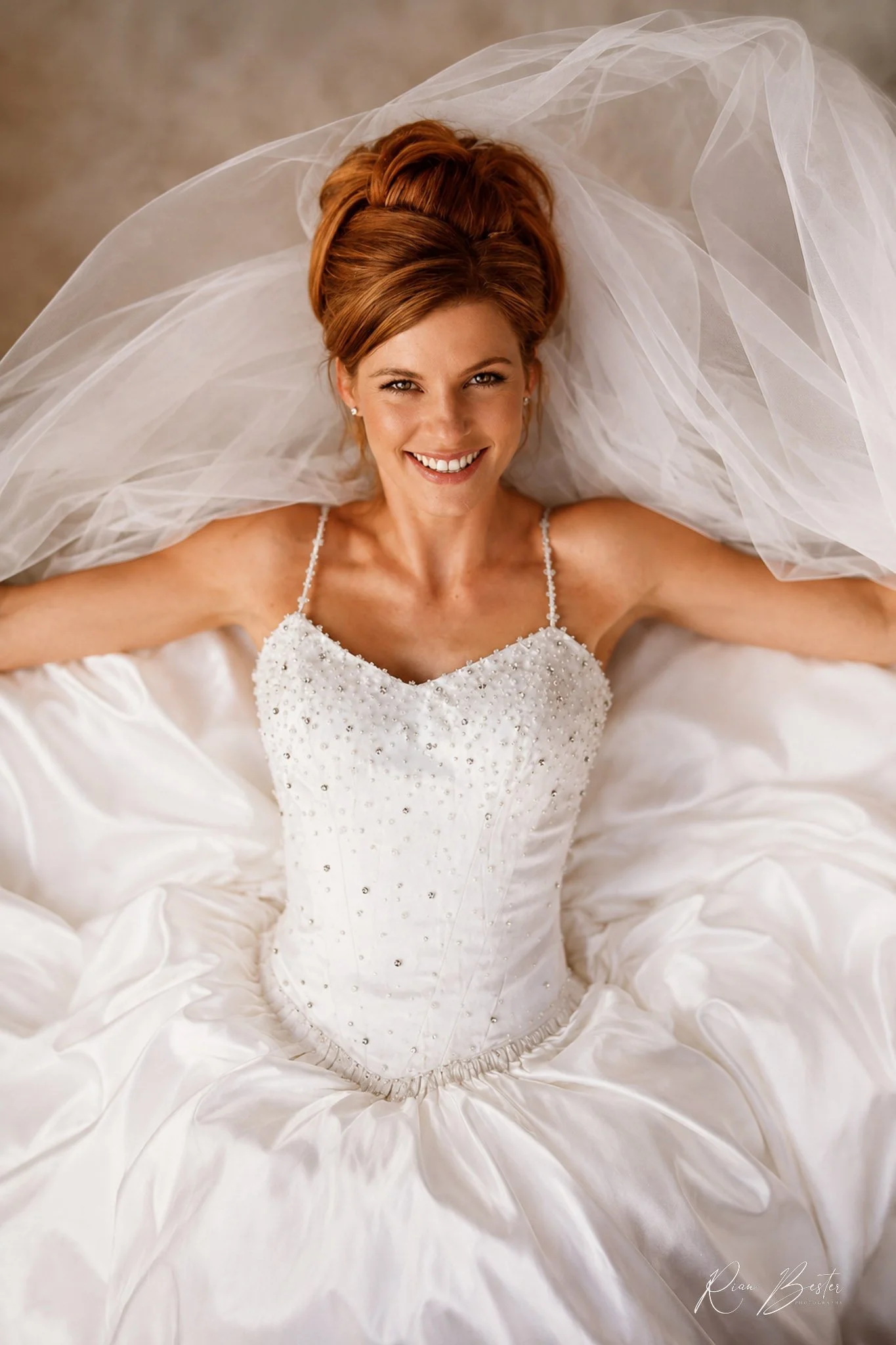 A smiling woman with red hair styled in an elegant updo, wearing a white wedding dress with beaded details, lying on a luxurious satin fabric with a sheer veil around her head.