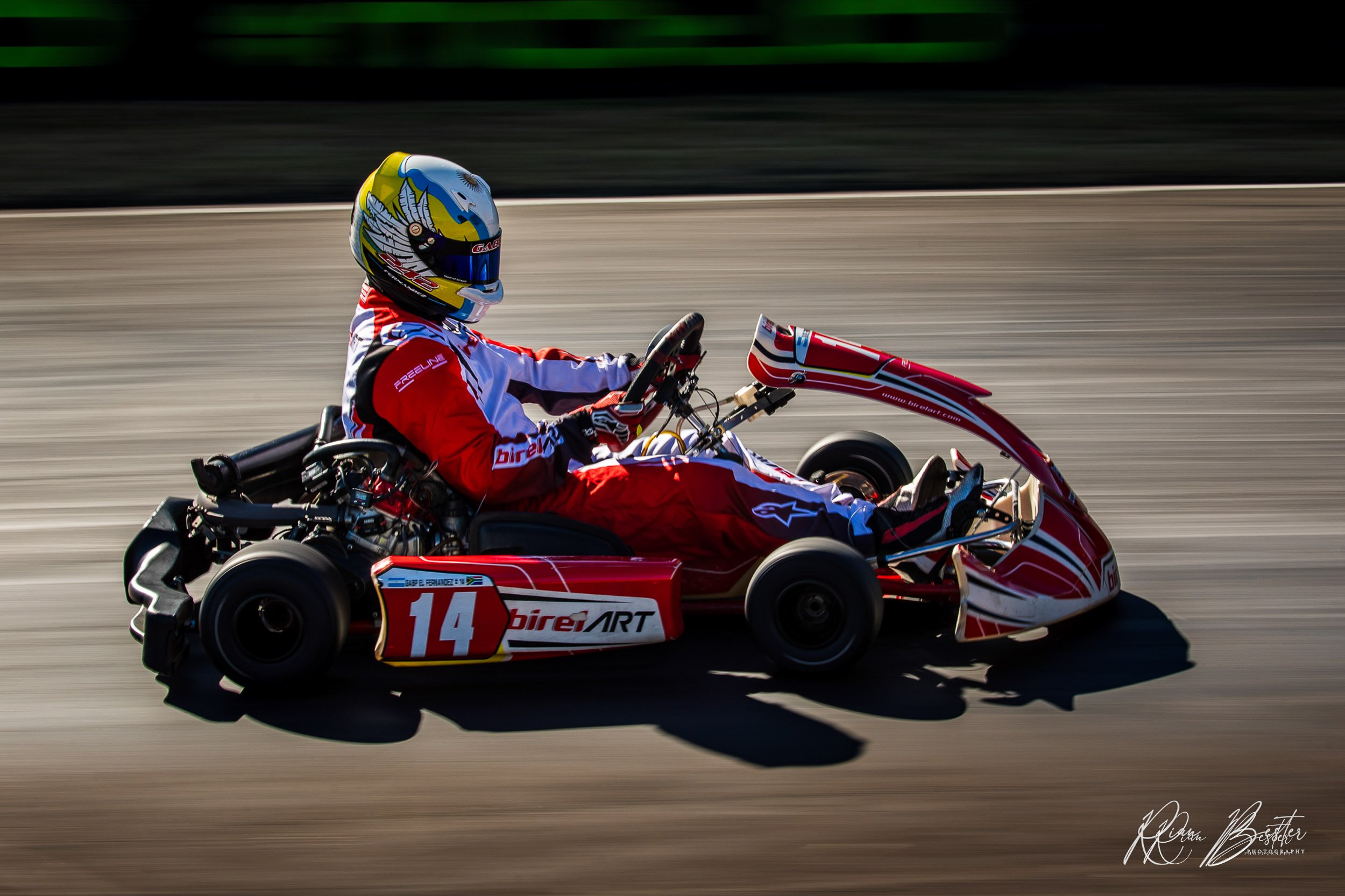 A go-kart driver in a red and white racing suit and blue helmet racing on a track
