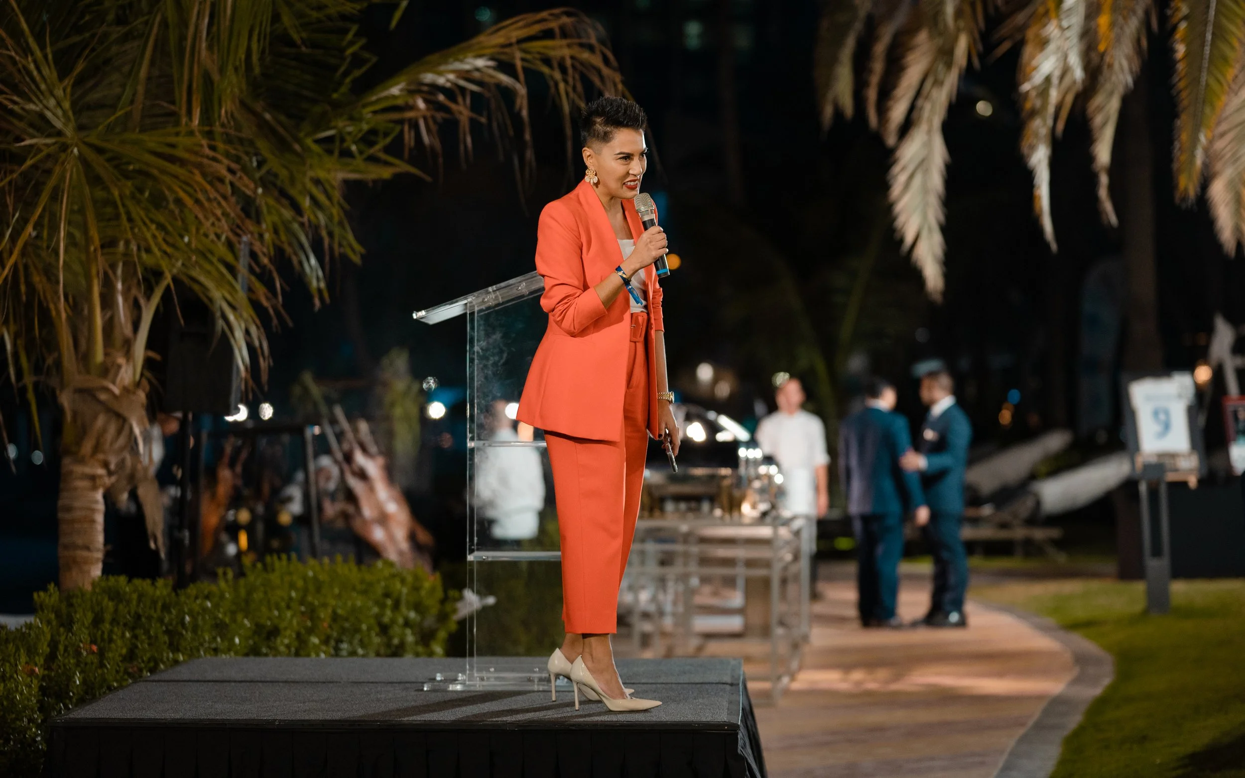 Sharon Pakir holding a microphone and addressing a large crowd on stage at an evening event in the UAE