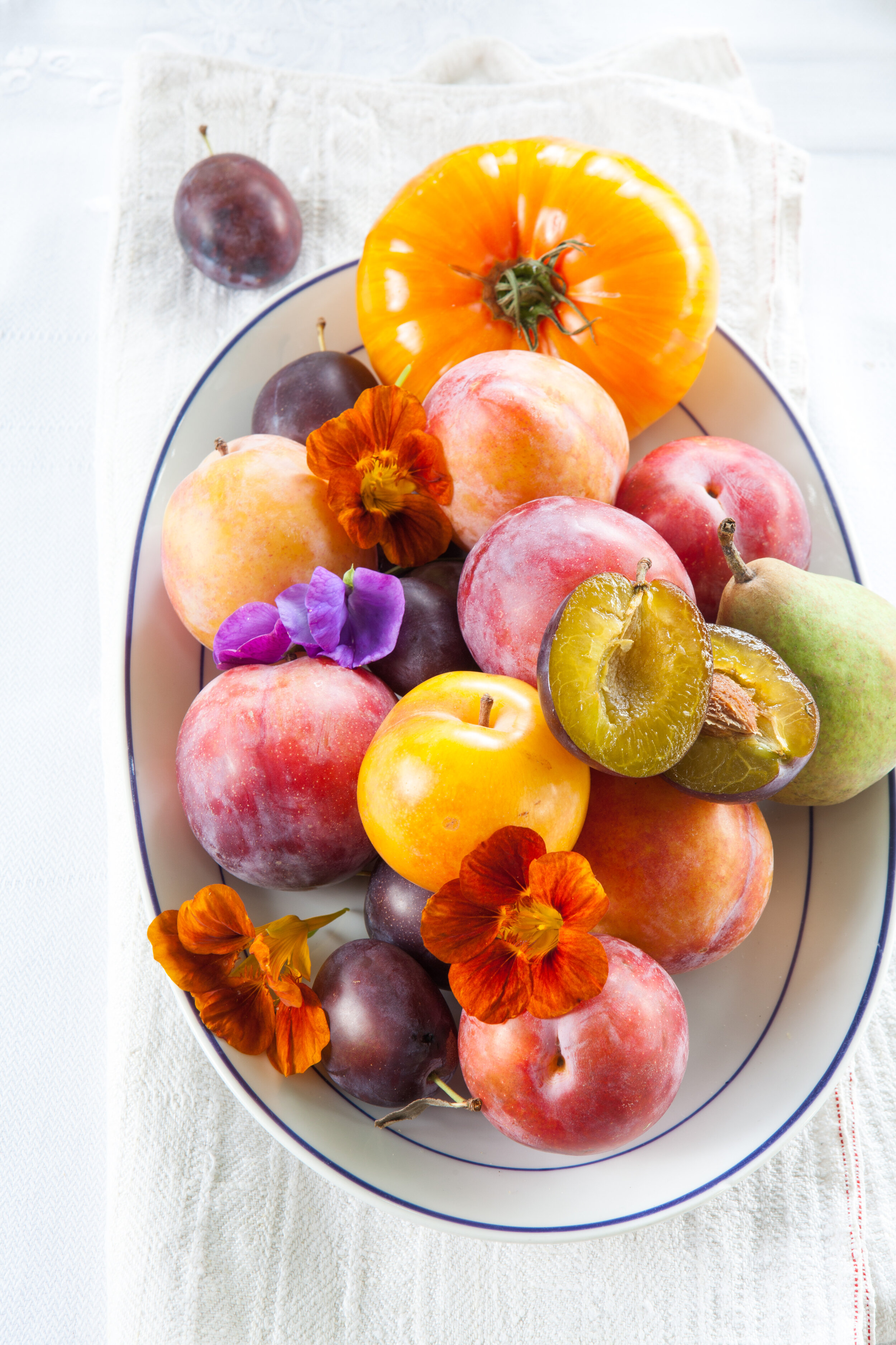 Een wit bord met een verscheidenheid aan fruit, waaronder pruimen, appel, perzik, nectarine, en een tomaat, versierd met oranje en paarse bloemen, geplaatst op een wit textiel met een rode stiksellijntje.