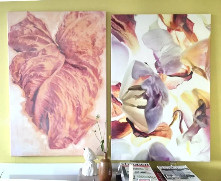 Twee grote schilderijen hangen aan een lichtgroene muur, één met een close-up van een bloemenblaadje en de andere met een close-up van veel bloemenblaadjes in verschillende kleuren, onder een venster. Hieronder staan een kleine witte beeldhouwwerk en