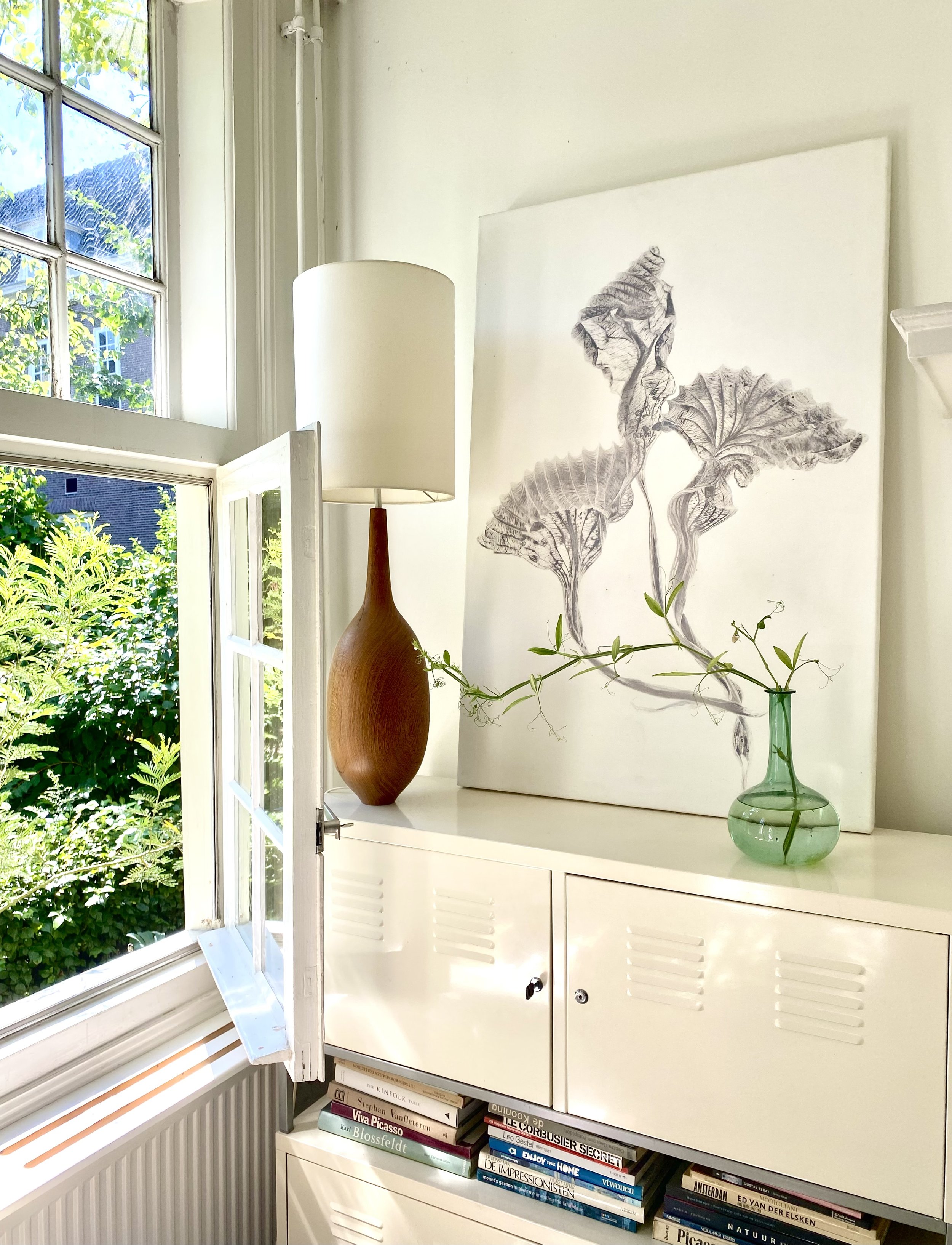 Binnenzicht van een witte kast met planken vol boeken onder een raam dat openstaat, met een grote lamp en een schilderij met gedetailleerd zwart-wit bladervorm