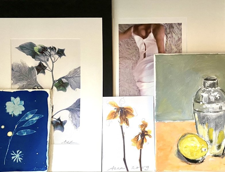 Een tafel met verschillende kunstwerken, waaronder aquarellen van bloemen, een foto van een vrouw in een witte jurk, en stilleven met een citroen en een potje.