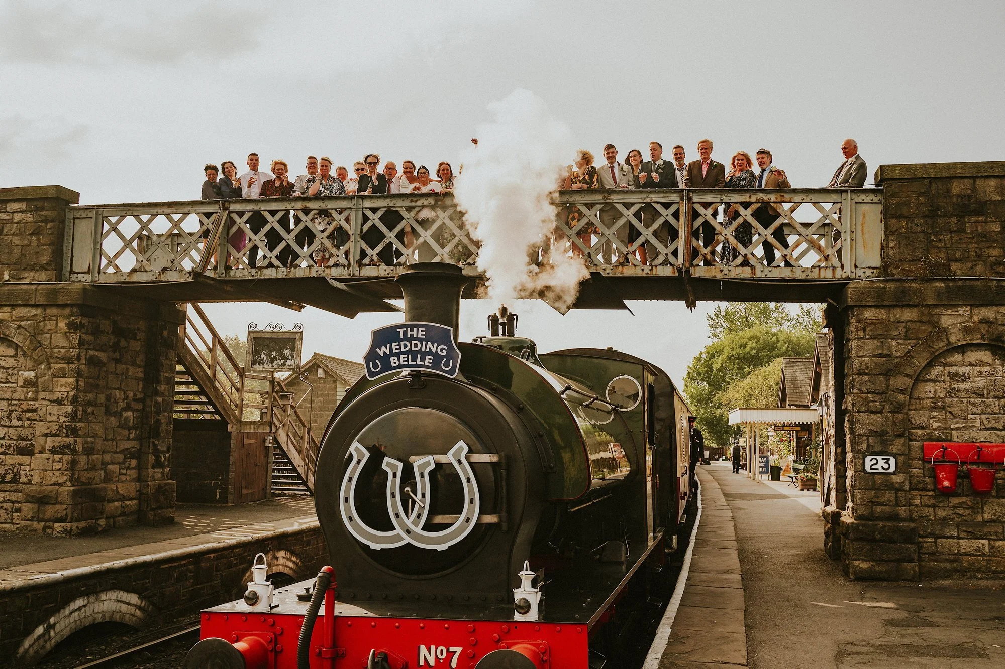 A group photo of the wedding party in this Embsay Bolton Abbey steam railway wedding. The group stand on a railway bridge while the train lets out a big puff of steam