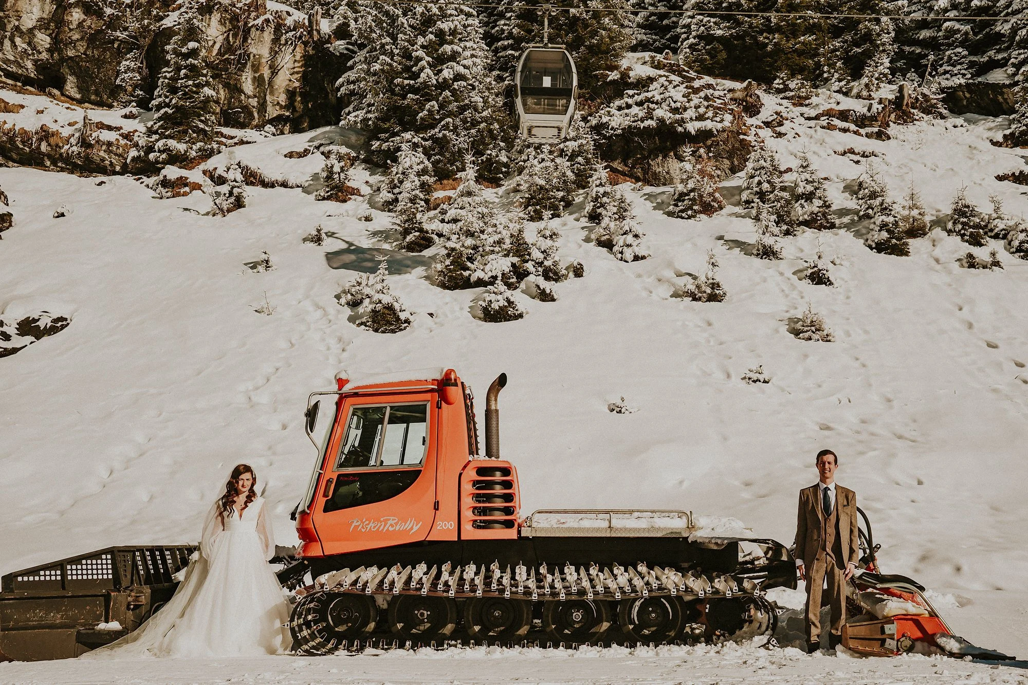 An orange snow mobile covered in snow provides a backdrop for a Bride and Groom just married at Lac Montriond. A gondola carrying skiers passes overhead.
