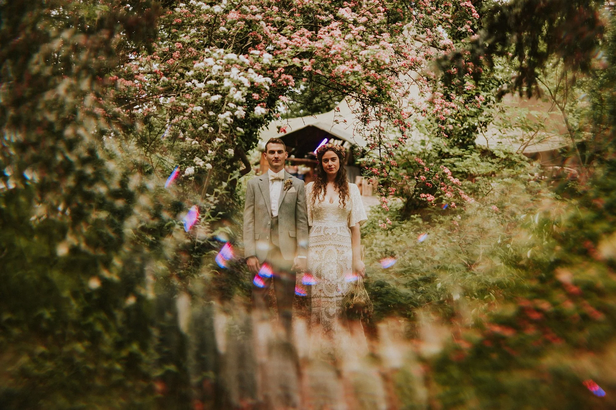 The Bride and Groom stand amidst blossom and beautiful fractal light amidst the woods in a This Green Moon wedding.