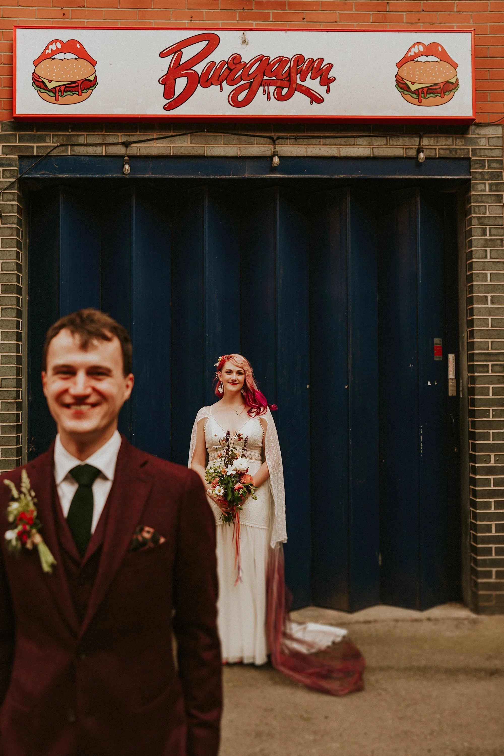 A Bride and groom pose outside the blue shutter of a burger restaurant in this Manchester urban wedding.