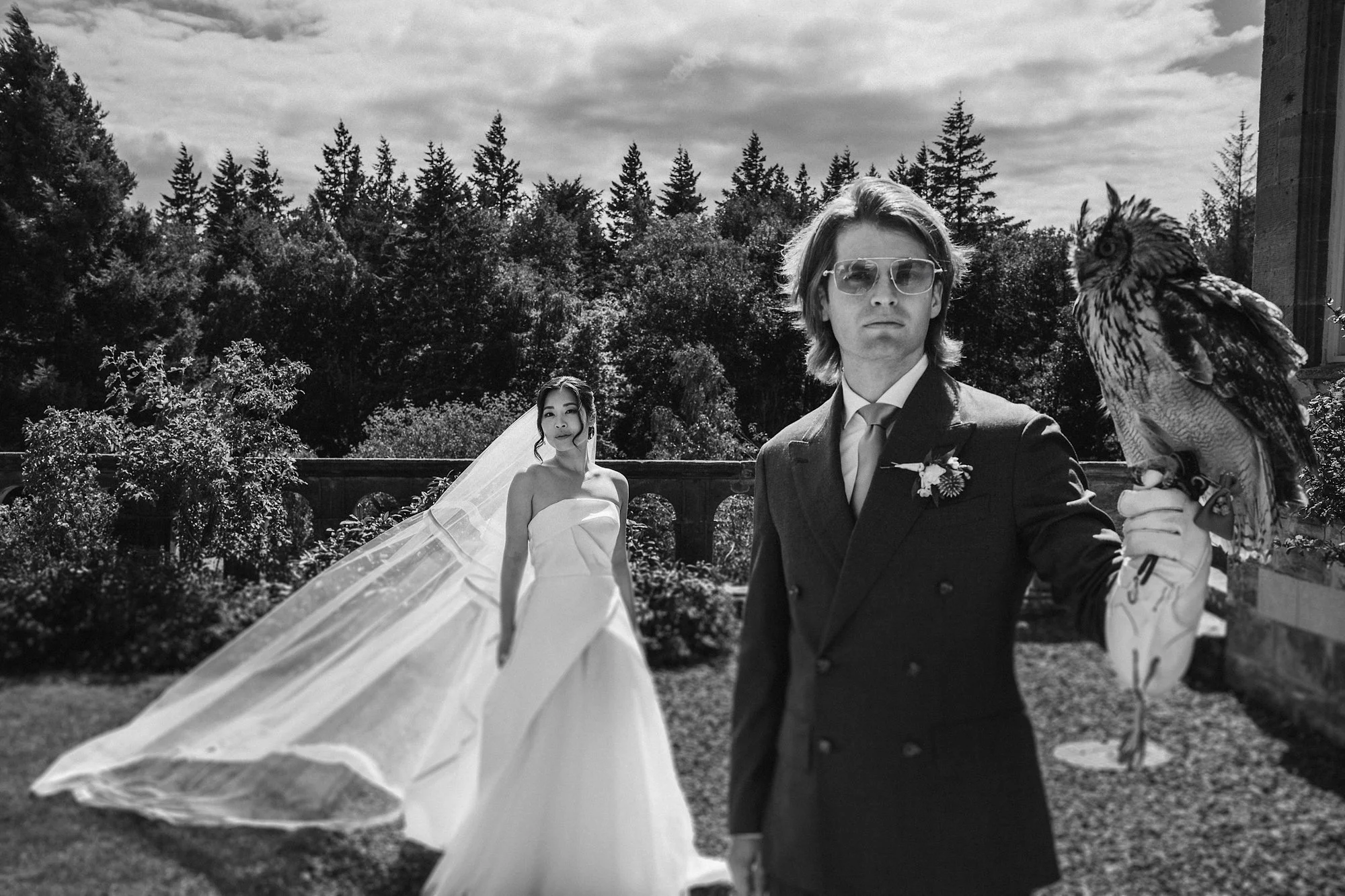 An atmospheric black and white portrait of a Bride and Groom. The Groom holds an owl while the brides veil cascades in Summer light.